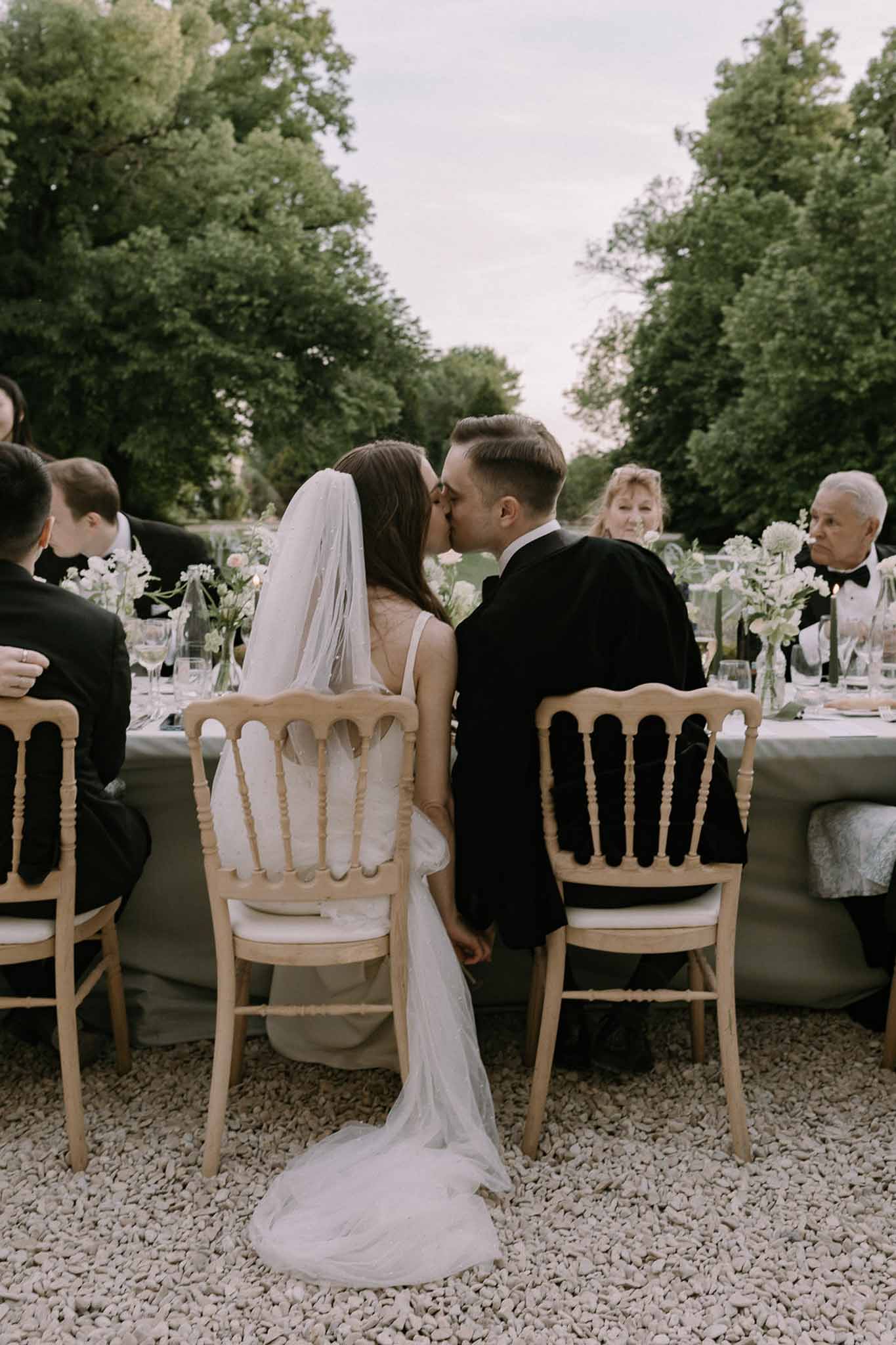 Bride and groom kissing at head table with bud vase florals and guests watching in garden reception