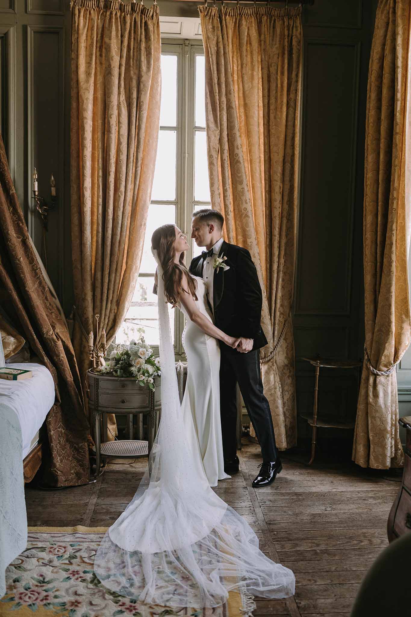Couple facing each other by gold-curtained window in olive-panelled room, bride's cathedral train trailing