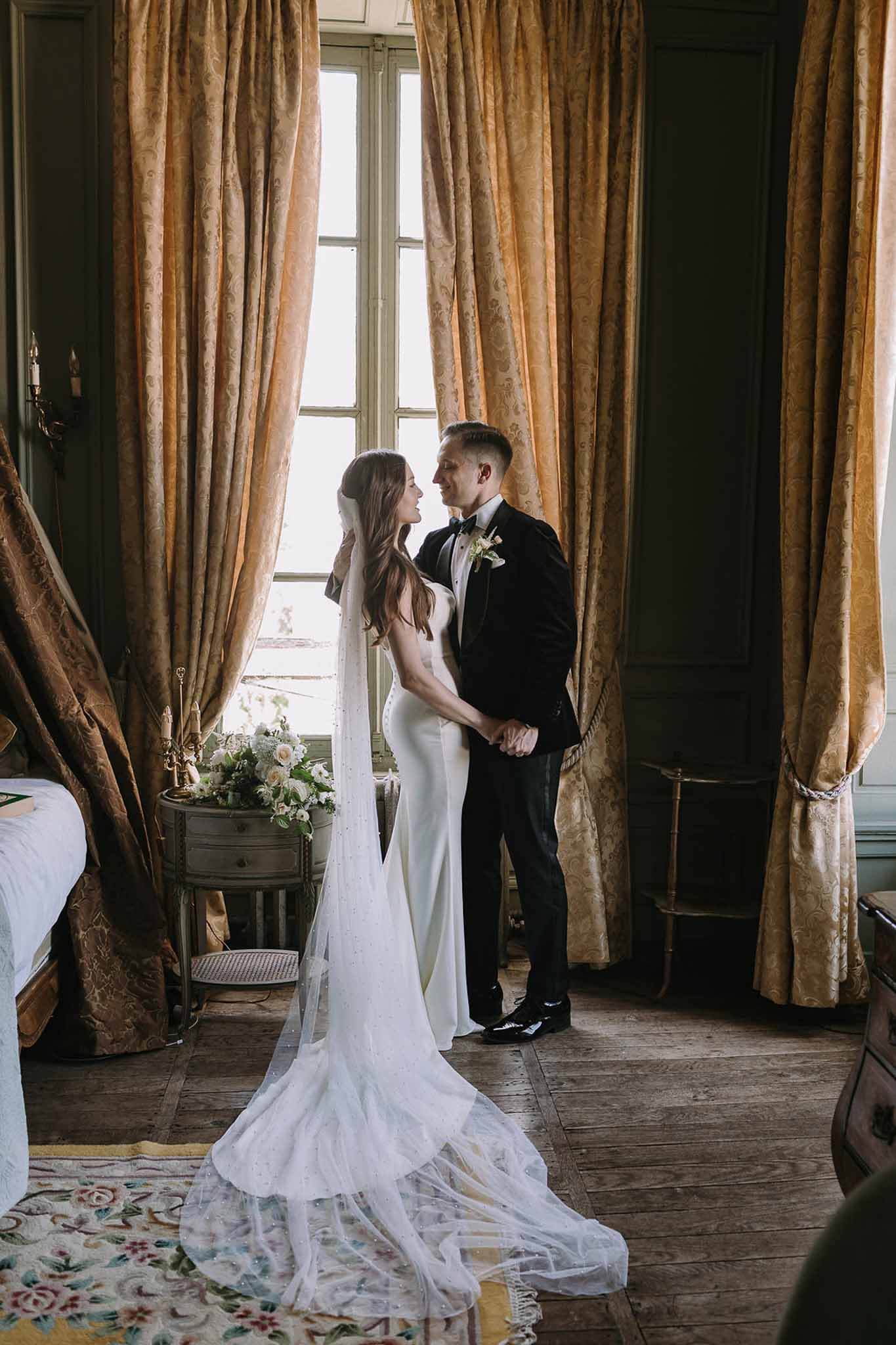 Couple holding hands in chateau salon with crystal-embellished veil trailing across antique rug