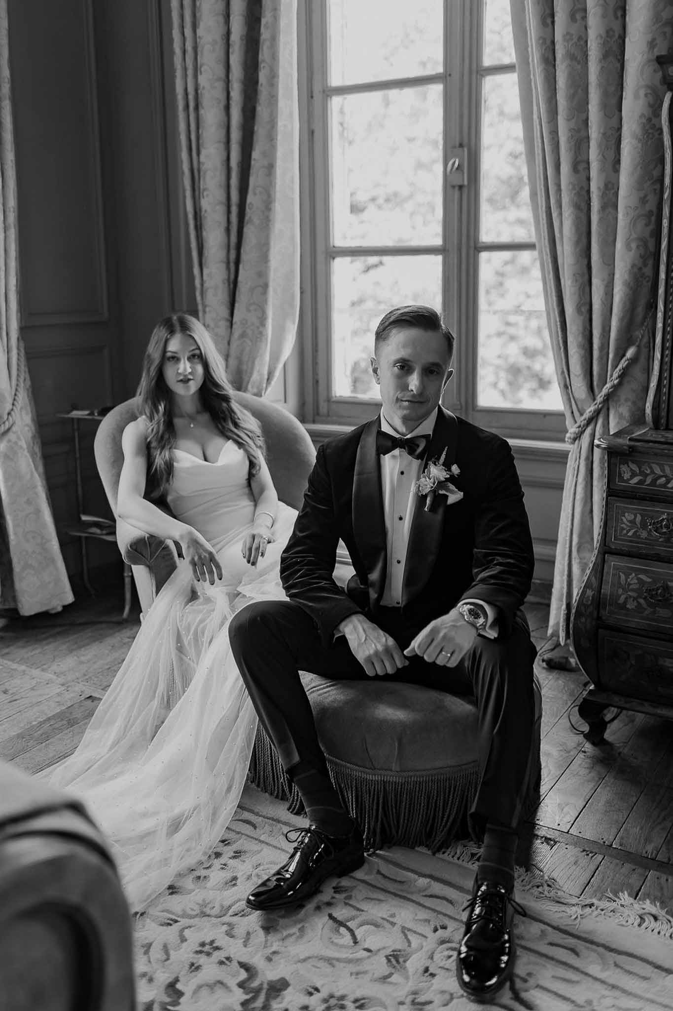 Black and white of couple seated in vintage chairs by tall curtained windows with ornate chest and parquet floor