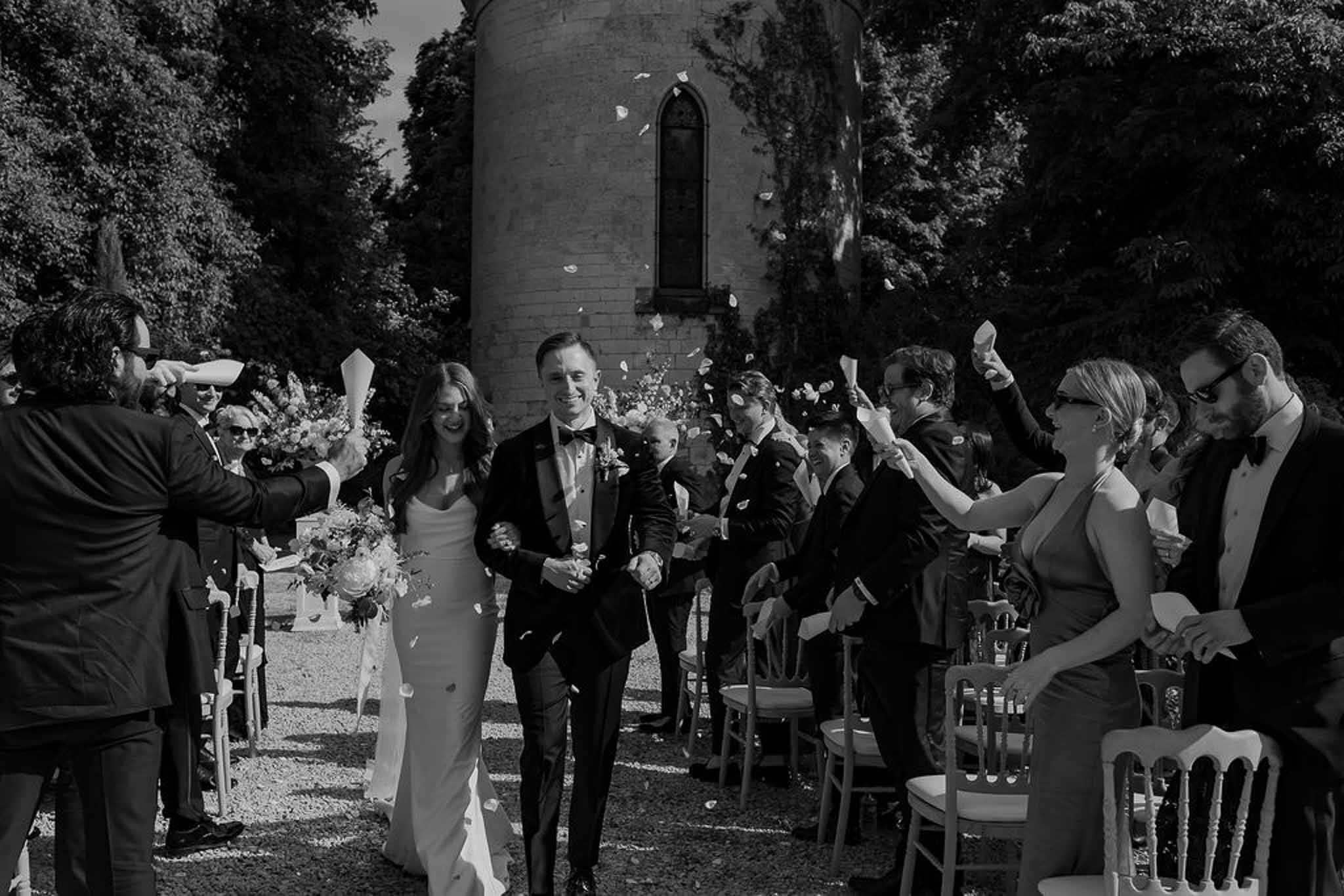 Black and white recessional with petal toss bride in slip gown with trailing bouquet guests and stone tower behind