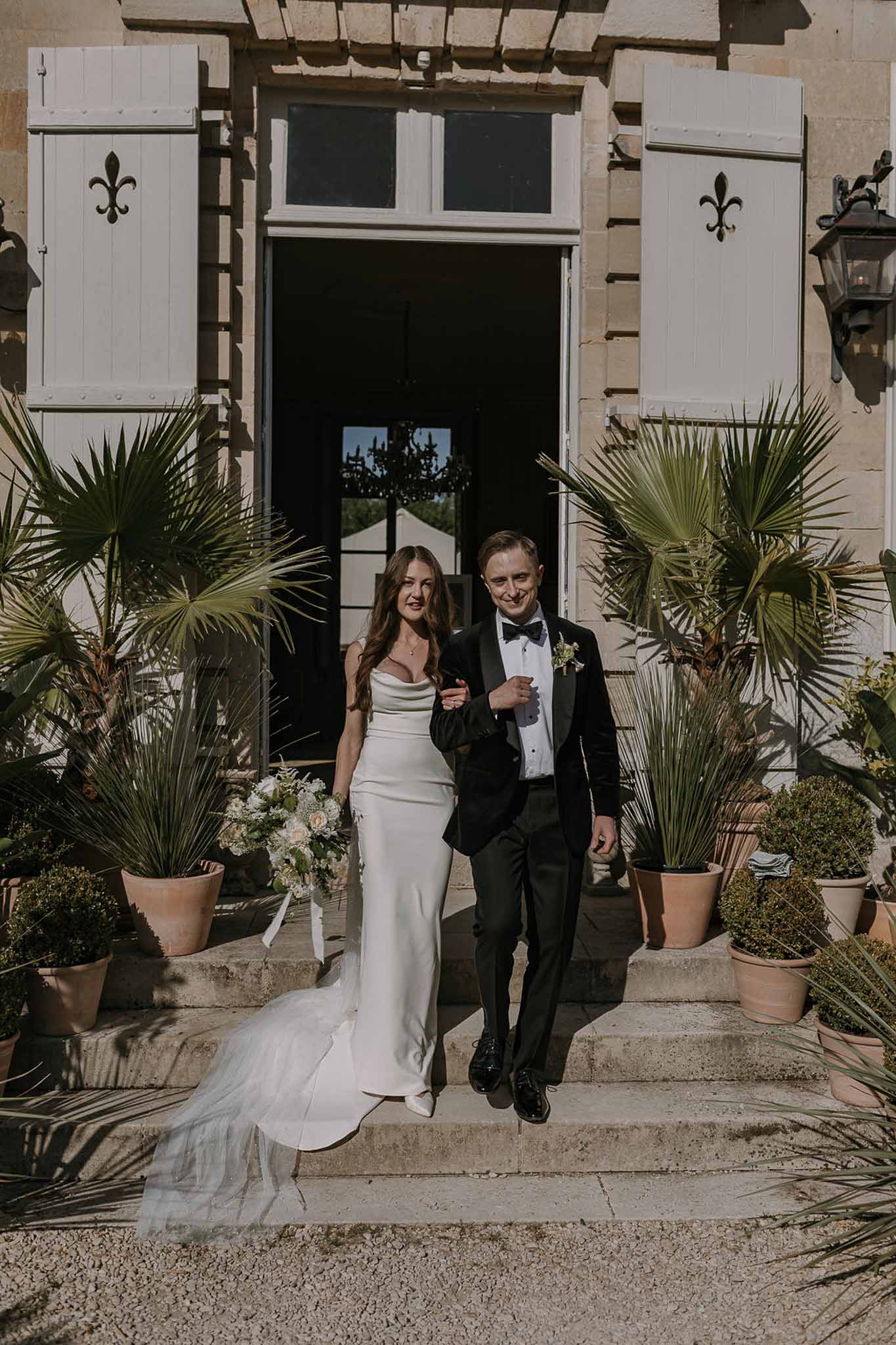 Couple walks down chateau entrance steps with palm trees and topiary holding trailing ivory bouquet