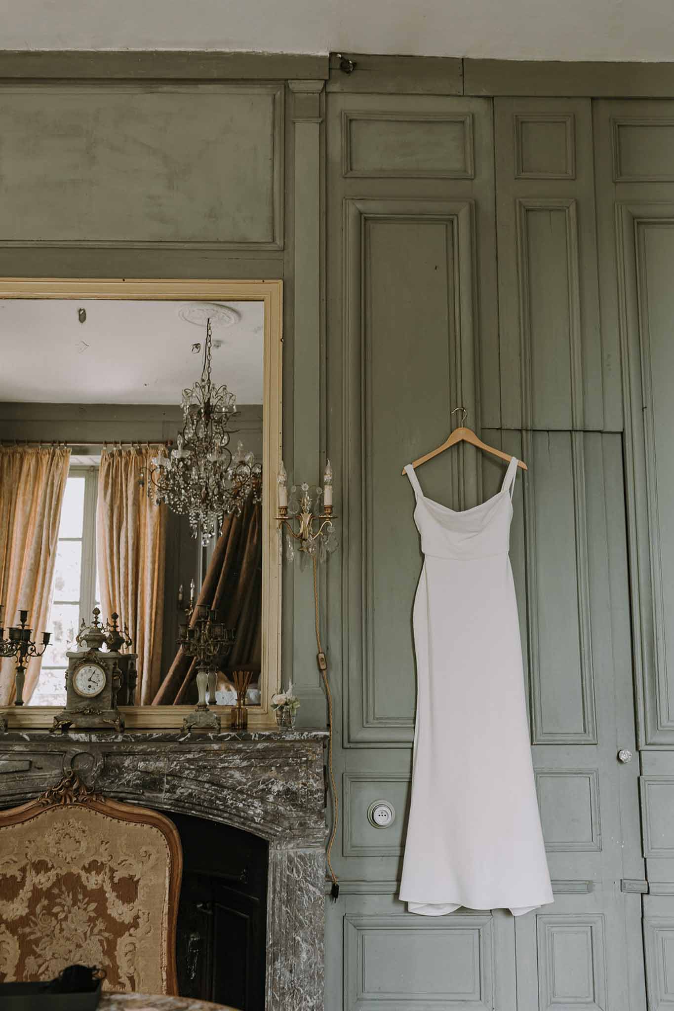 Ivory slip wedding dress on hanger beside grey marble fireplace with gilt mirror in sage-green chateau bedroom