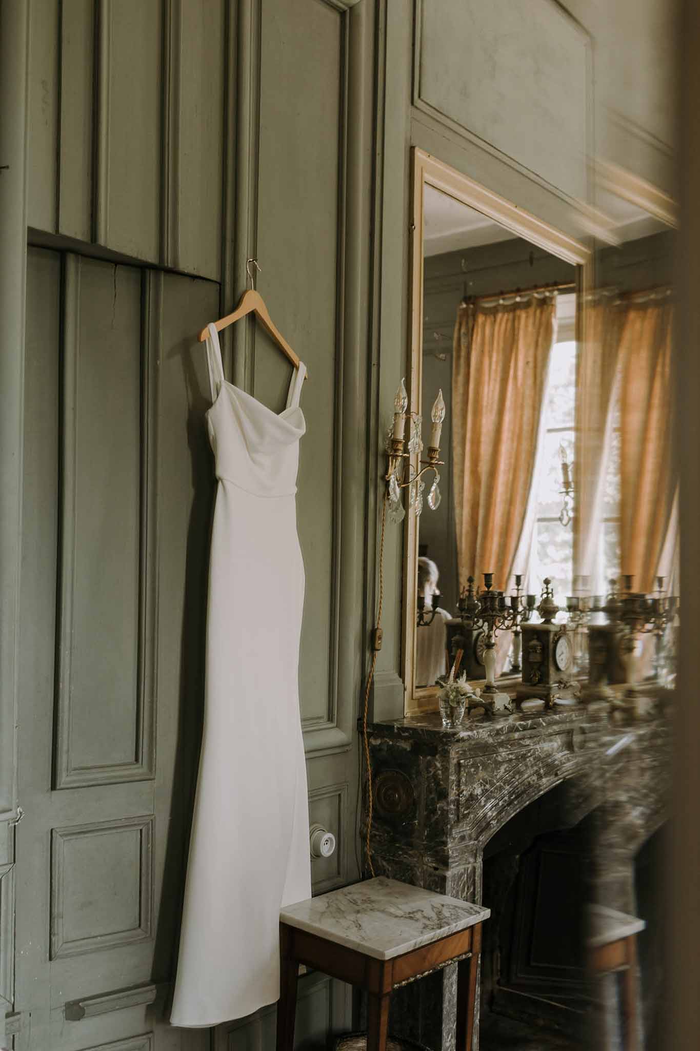 Ivory slip dress hanging on sage green panelling beside marble fireplace with brass candelabras
