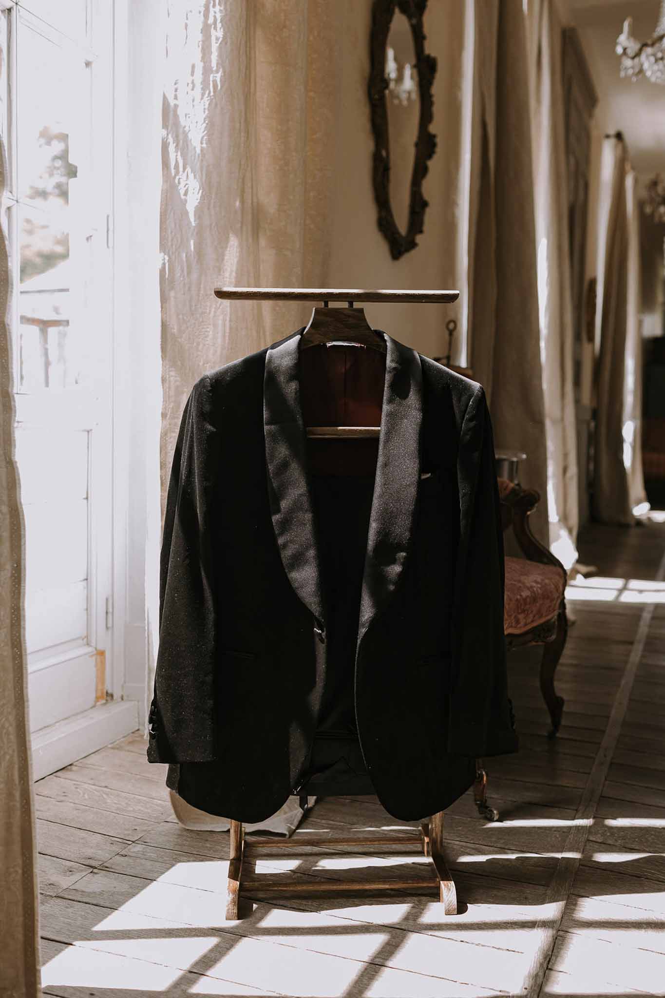 Black tuxedo jacket on valet stand in chateau room with chandelier and window-pane shadows