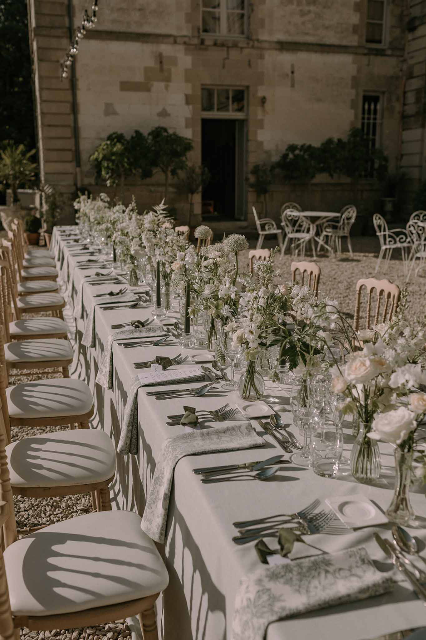 Long feasting table with botanical napkins, white ranunculus bud vases, and black taper candles in chateau courtyard