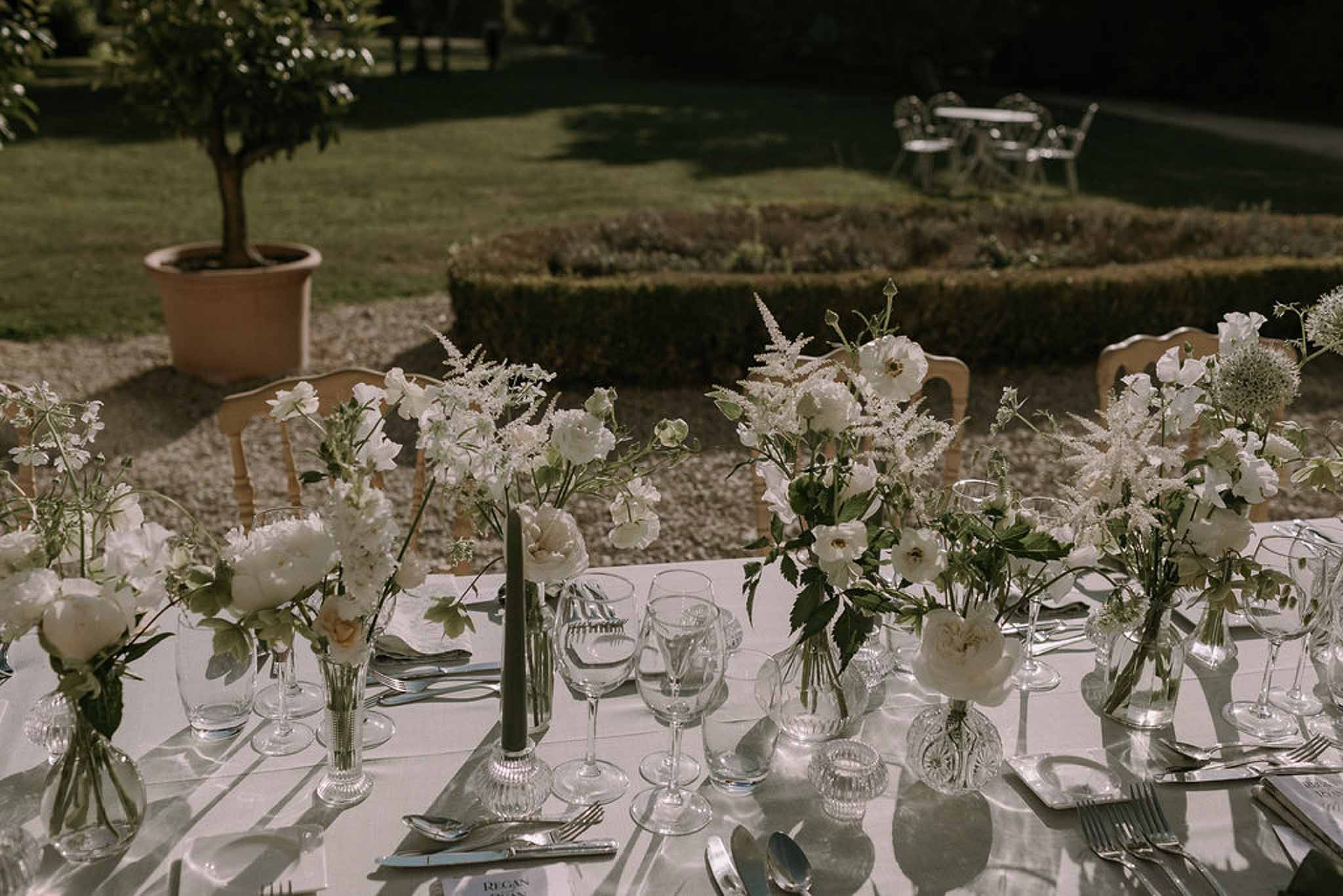 Round table with white peony bud vases, dark green tapers, and crystal tumblers in formal garden setting