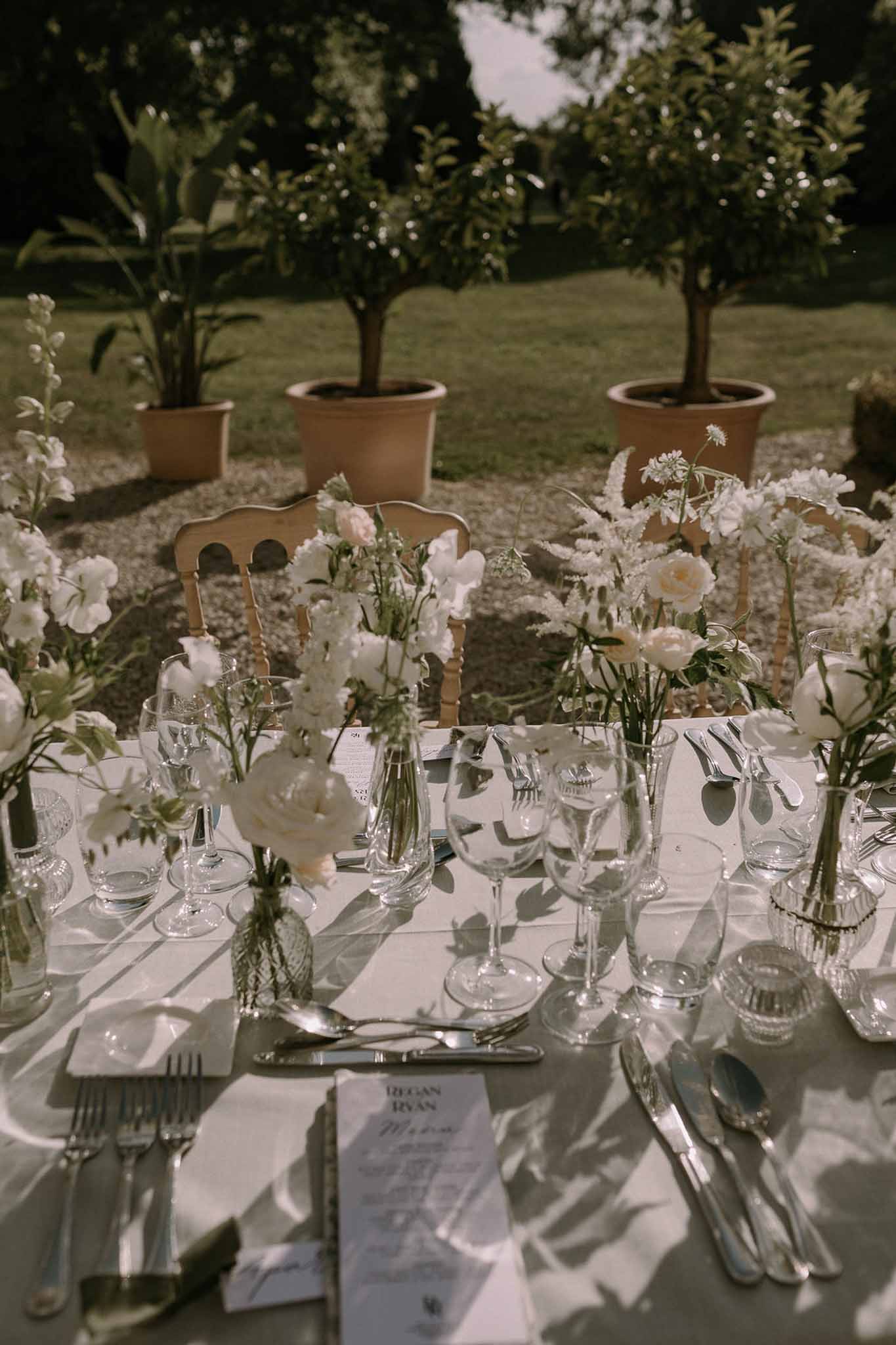 Garden reception table with white bud vases of roses and ranunculus, printed menu cards, and crystal glasses
