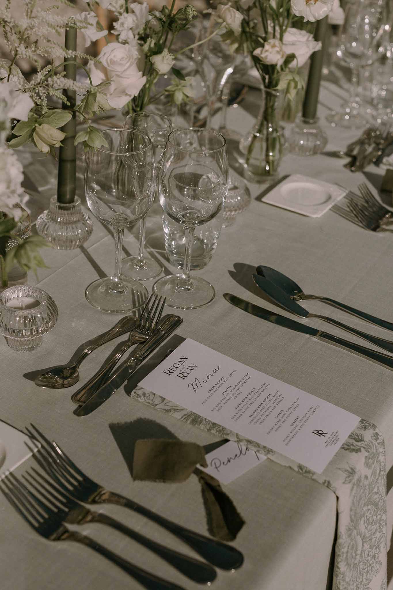 Wedding table setting with sage green linen, crystal glasses, white roses, menu card, and forest green taper candles