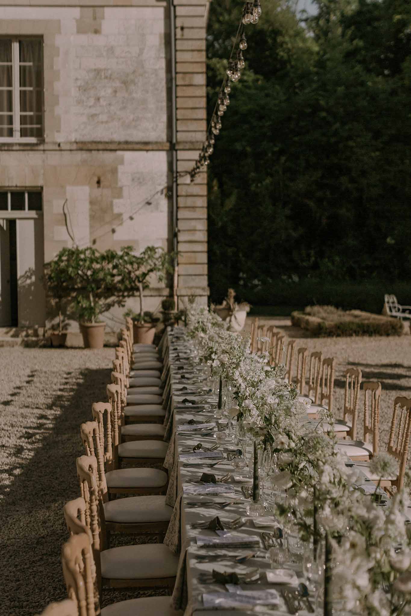 Long feasting table with gold Napoleon chairs and white floral centerpieces under string lights in chateau courtyard