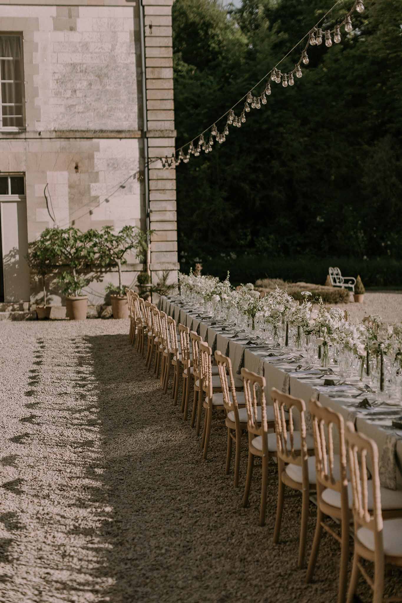 Long banquet table with bud vases and Edison lights on gravel courtyard beside pale stone chateau