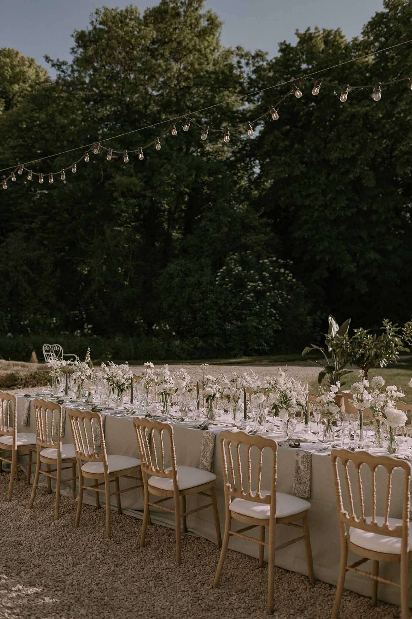 Banquet table with white ranunculus bud vases, dark tapers, and Napoleon chairs under Edison lights