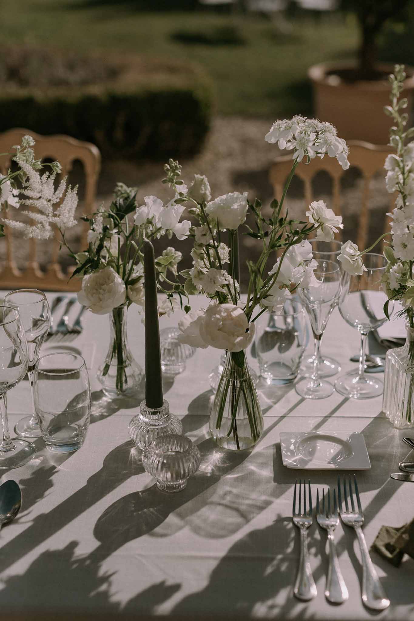 White peony and sweet pea bud vases with dark green taper candle and silver cutlery in sunlit garden