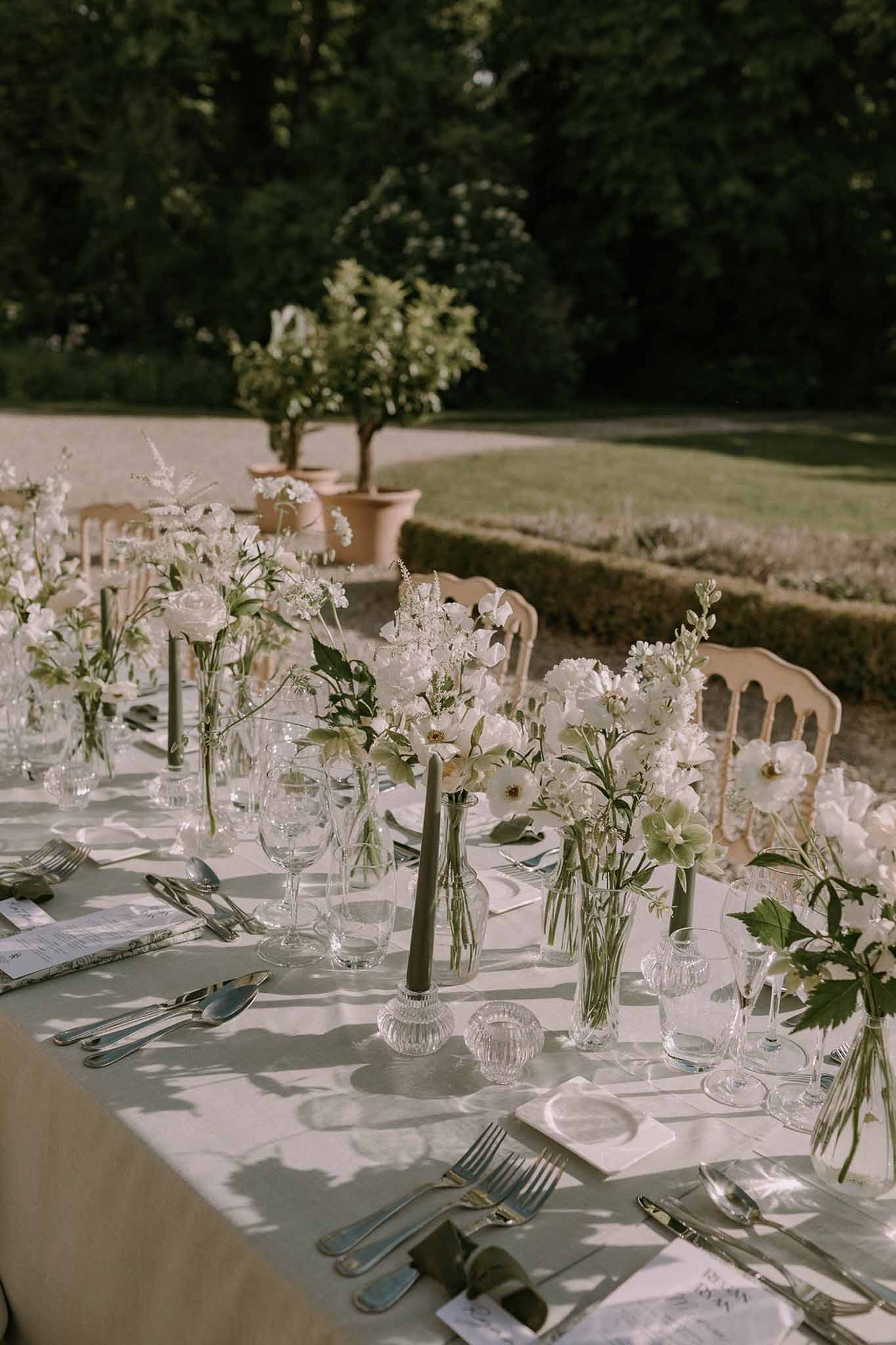 White anemone and sweet pea bud vases with olive taper candles and silver flatware in formal garden
