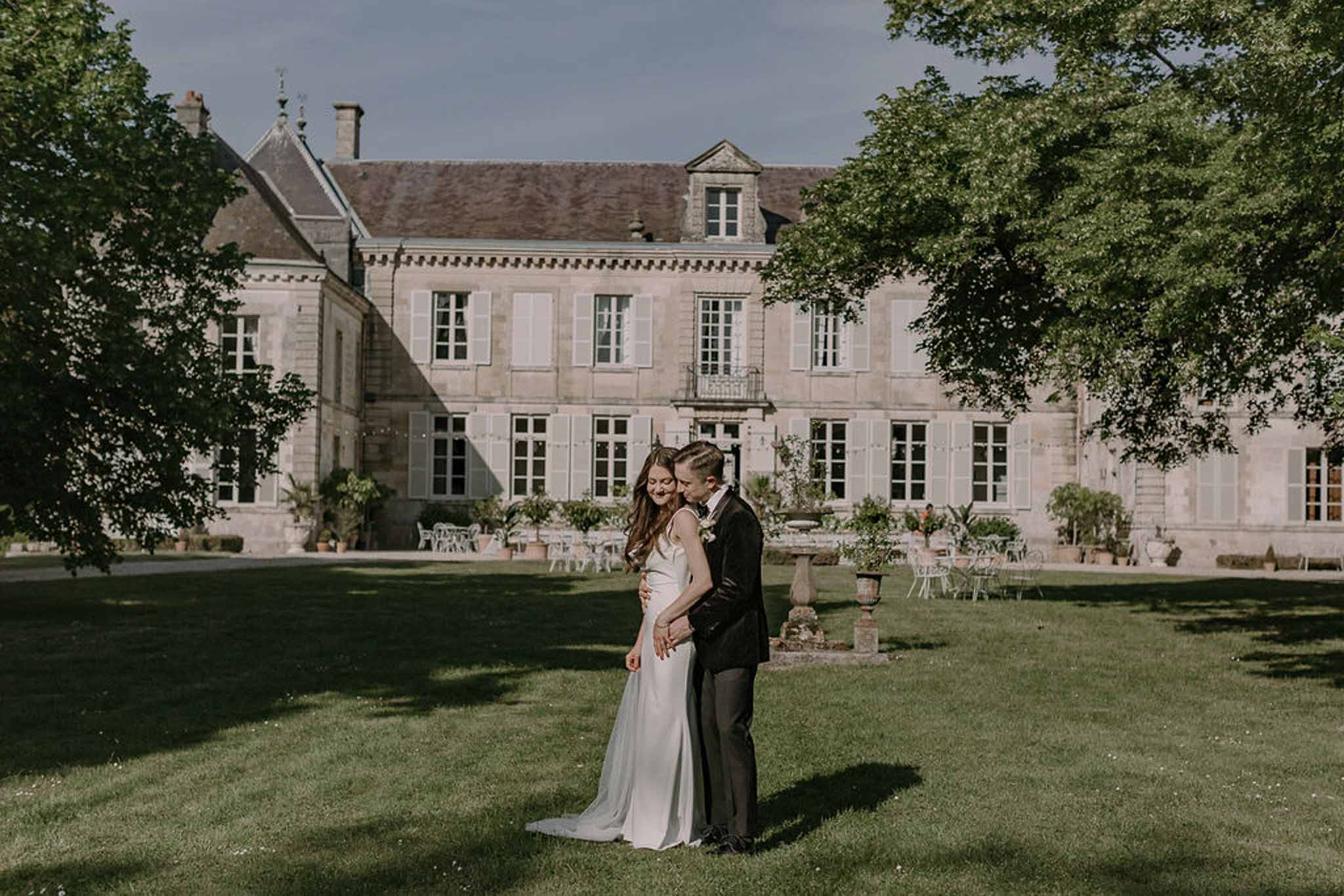 Bride and groom on chateau lawn with full stone manor facade and garden furniture in background