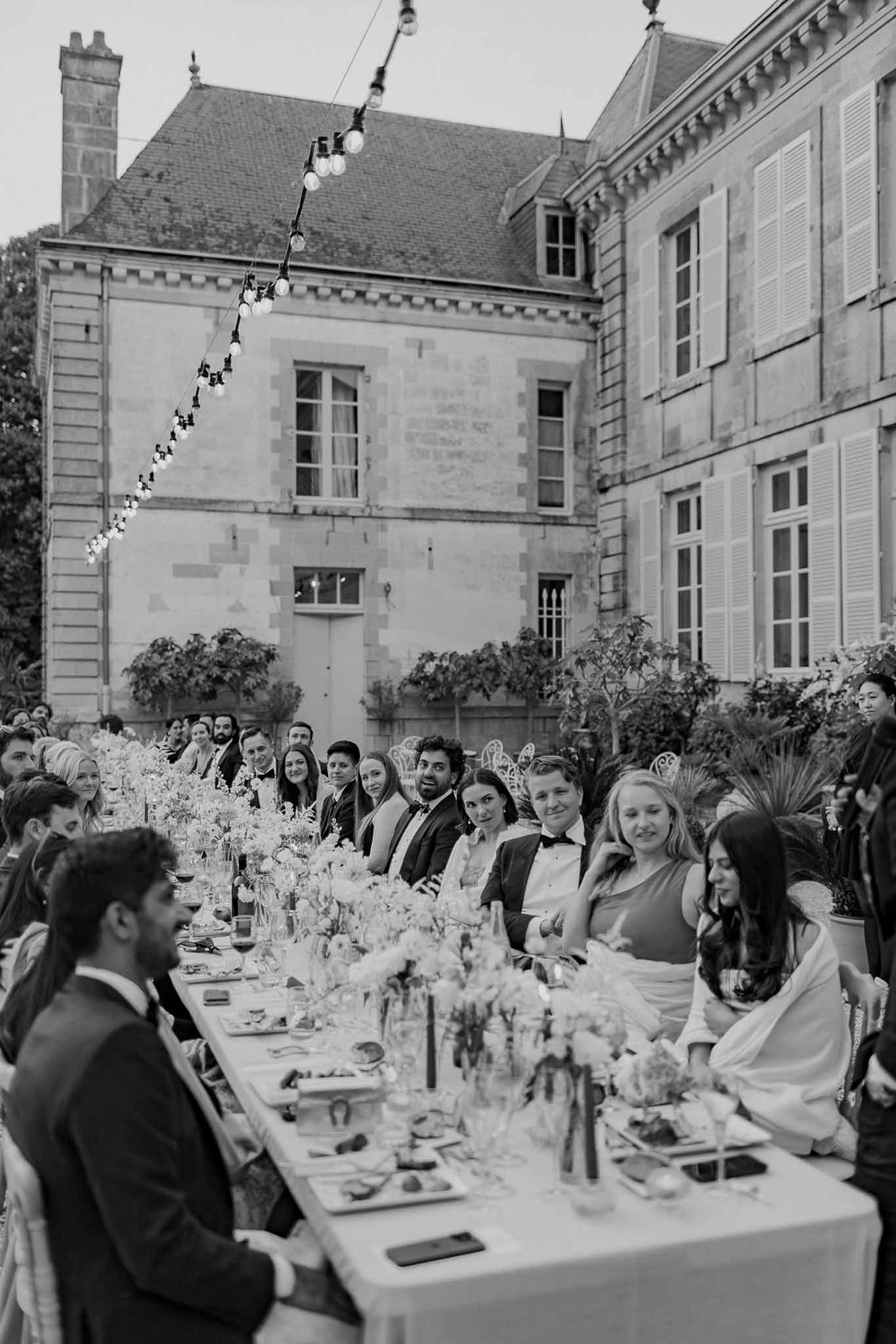Black and white reception dinner with 30 guests at long table with Edison lights in chateau courtyard