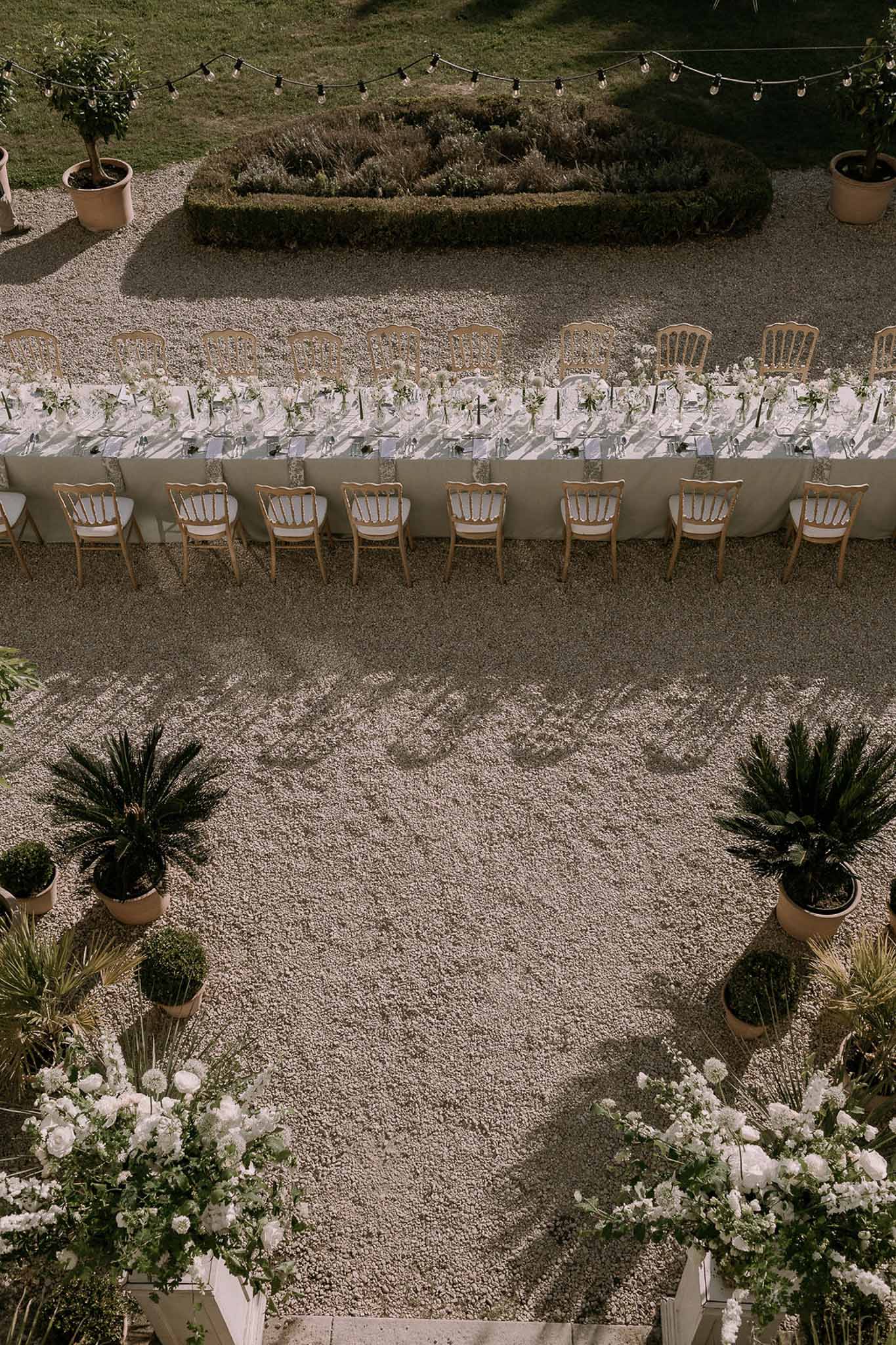Aerial long feasting table with white ranunculus clusters and festoon lights flanked by palm planters on gravel courtyard