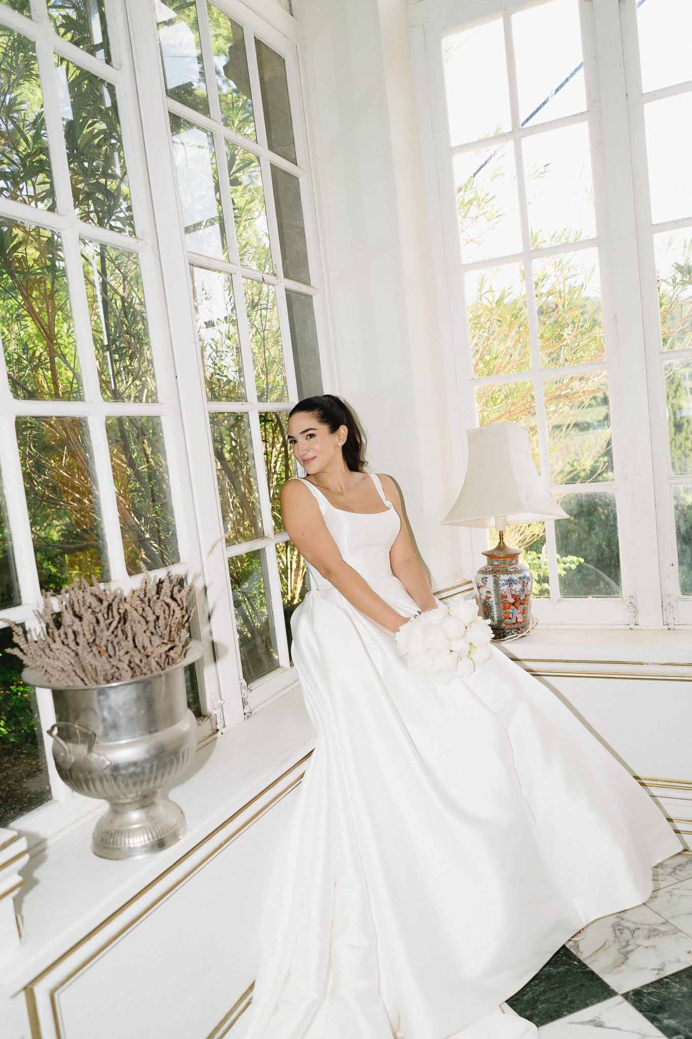 Bride in white satin gown seated on console table in chateau interior with checkered marble floor