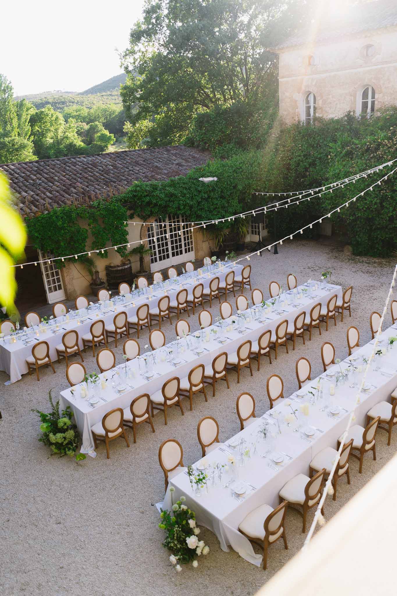 Aerial view of four long banquet tables with white linens and bistro lights in a Provencal stone courtyard