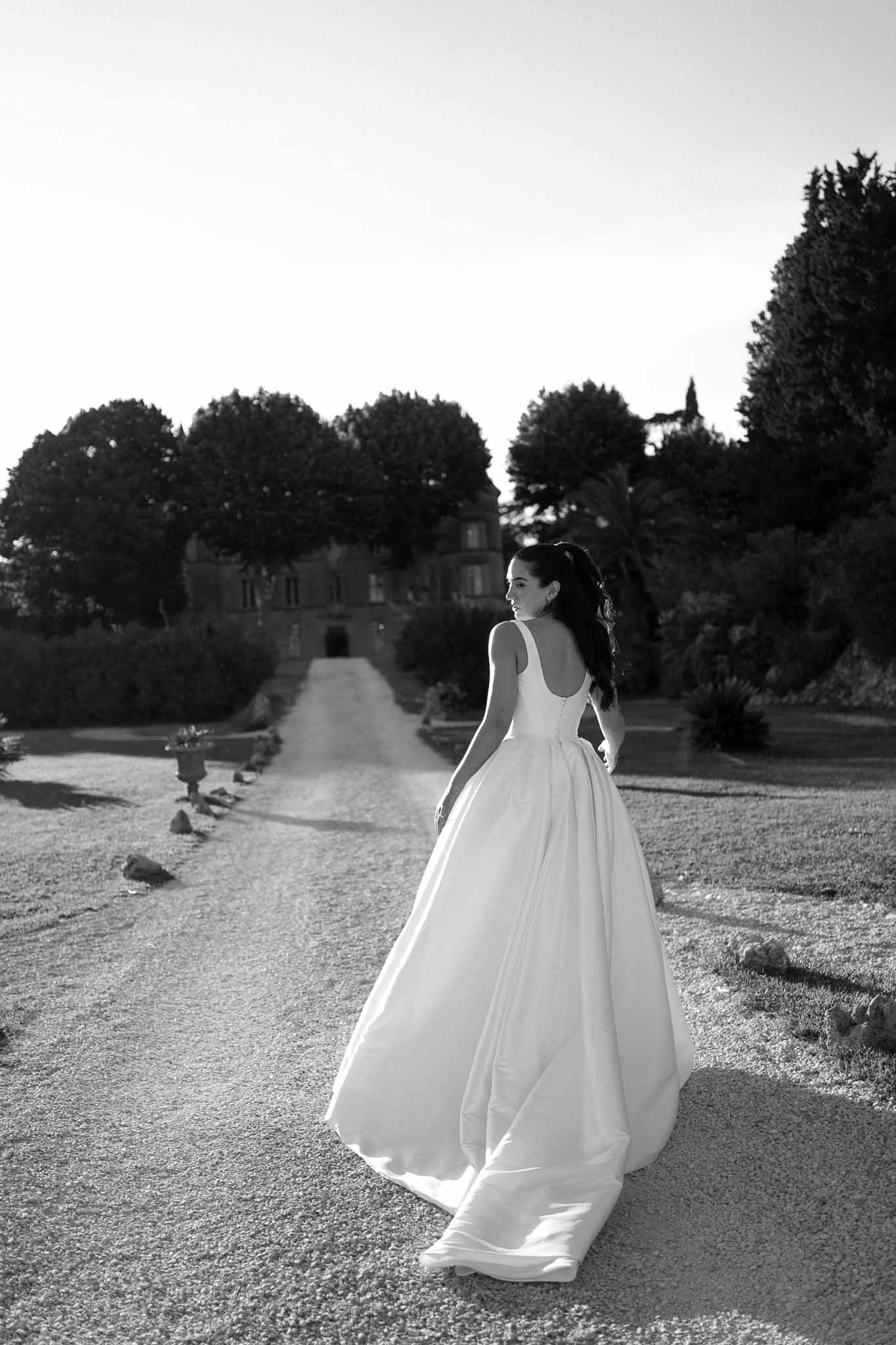 Black and white portrait of bride on chateau gravel driveway, ball gown train spread across ground, glancing over shoulder