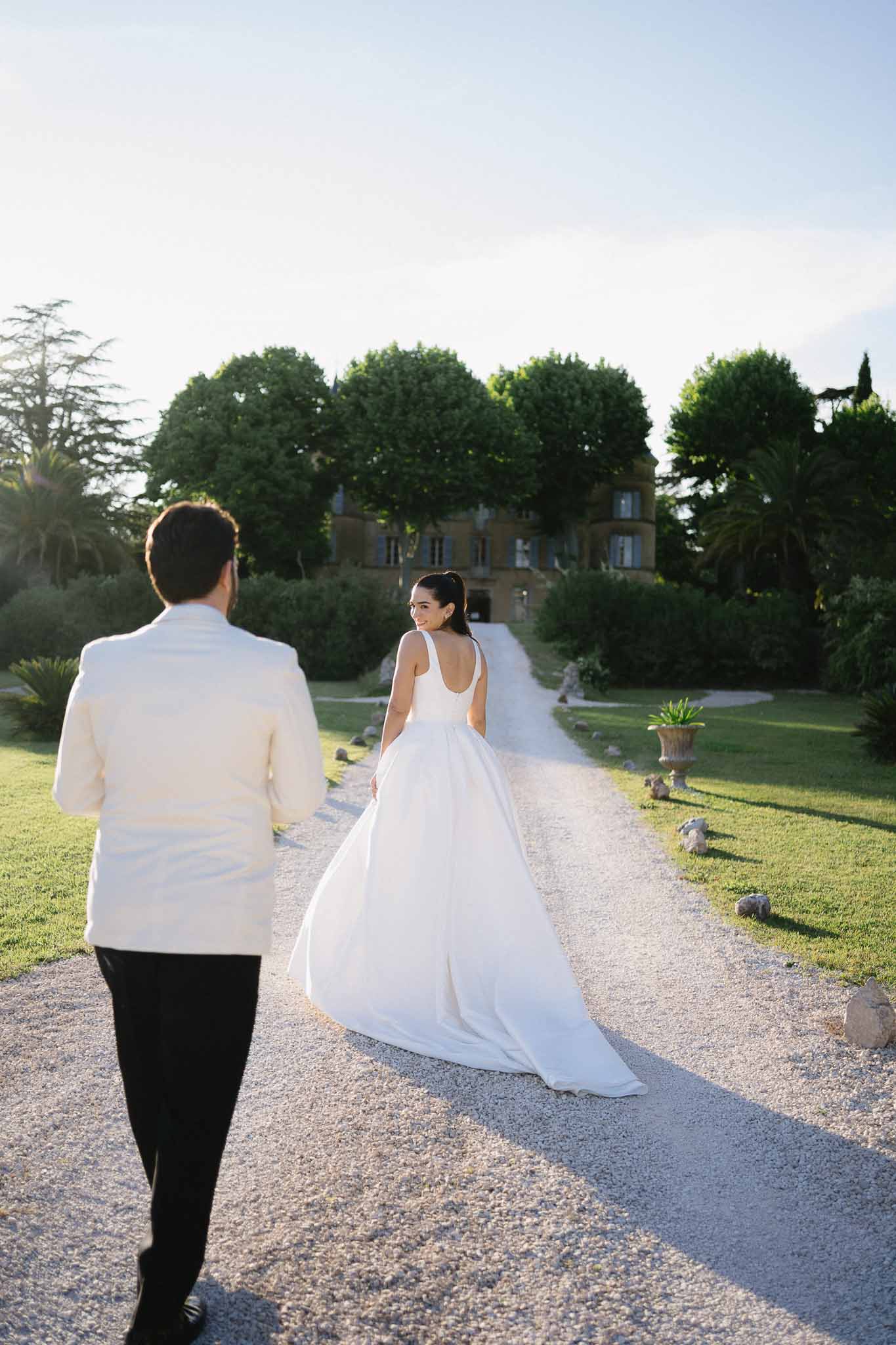 Couple from behind walking toward honey stone chateau bride glancing back in scoop-back gown groom in ivory dinner jacket