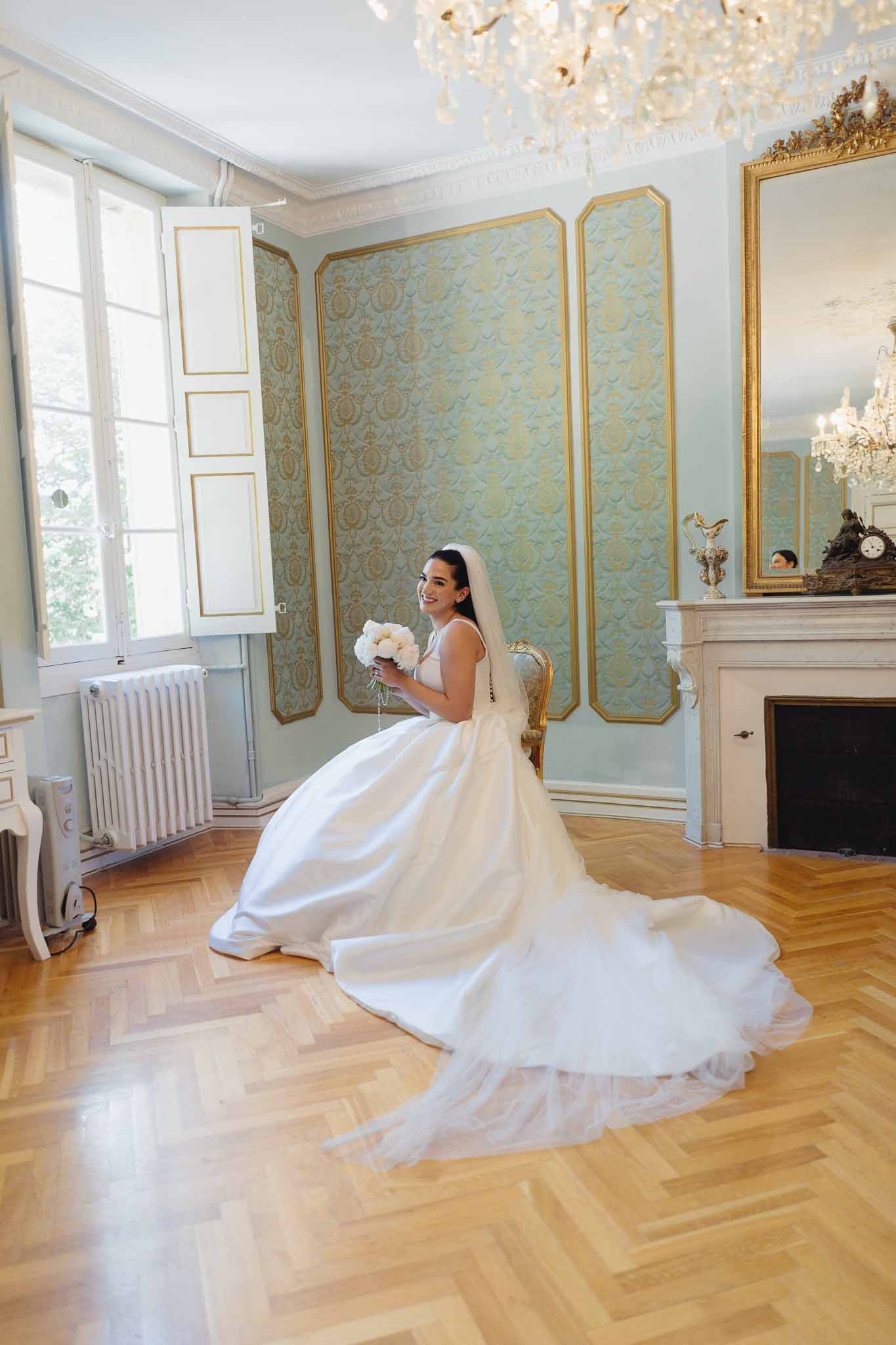 Bride seated in ornate chateau salon wearing white ball gown with cathedral veil holding white peony bouquet