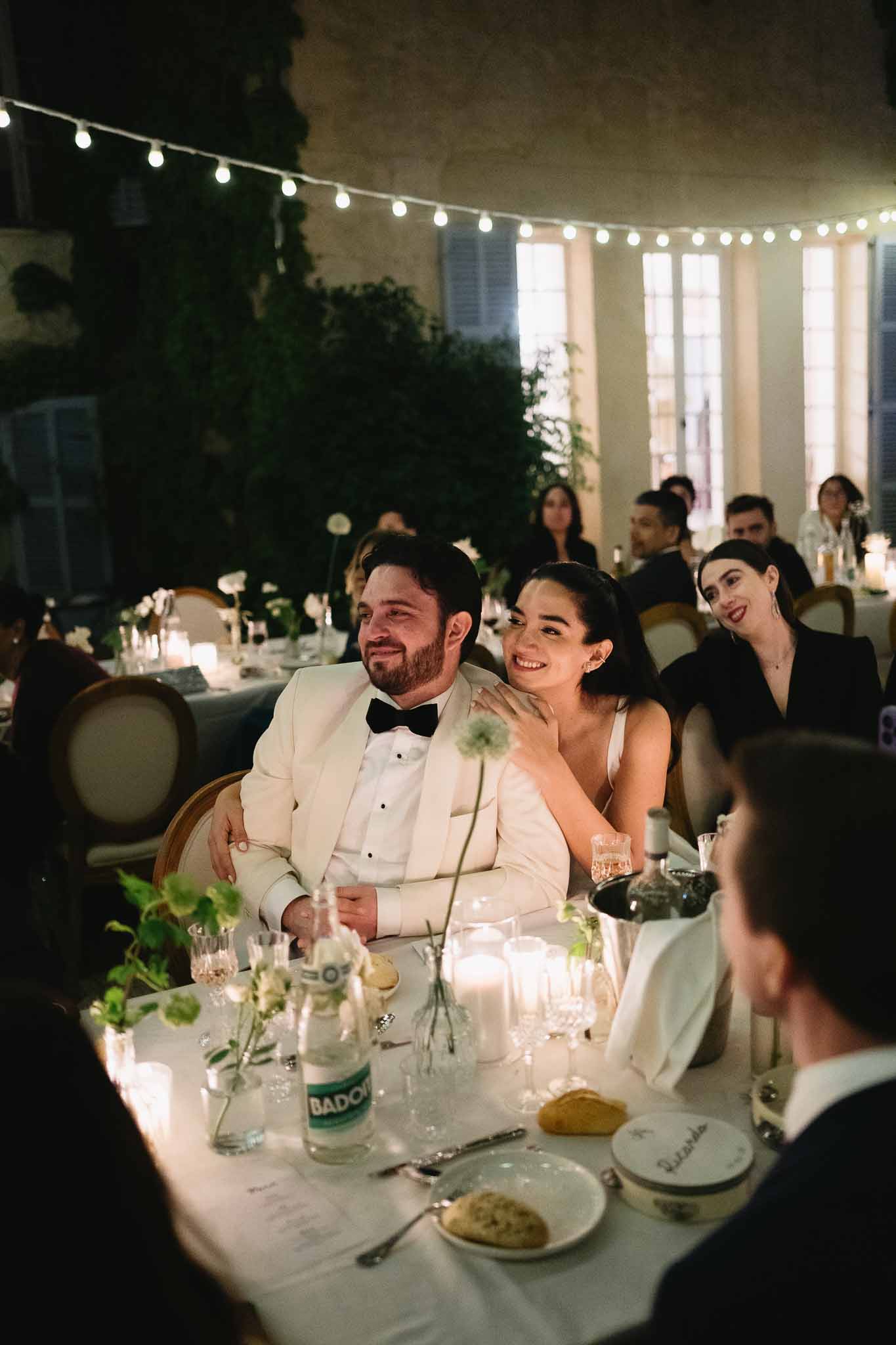 Bride and groom seated at sweetheart table during reception speeches under string lights in a stone courtyard
