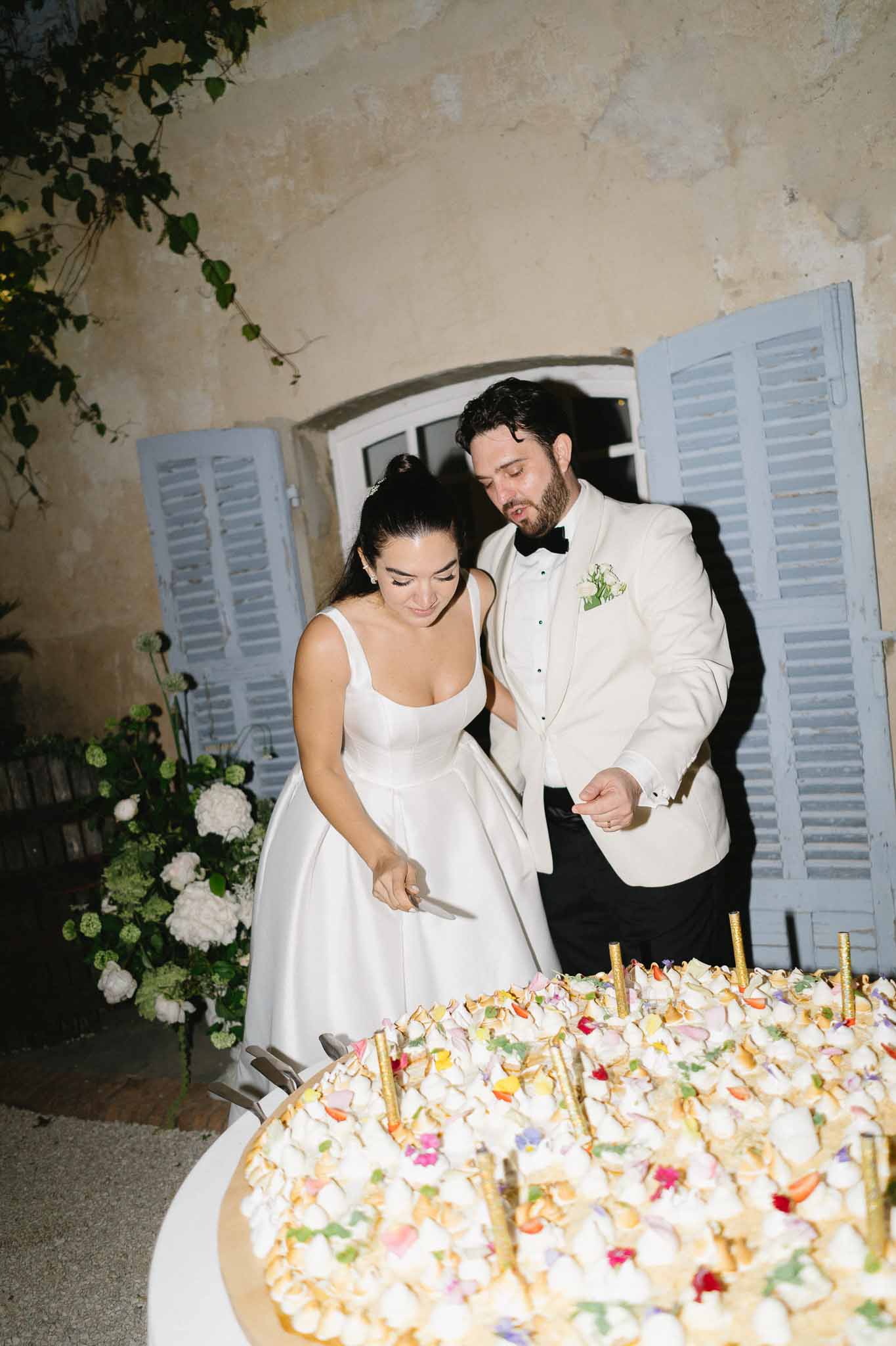 Couple cutting tart-style cake with edible flowers and gold candles beside white hydrangea at night