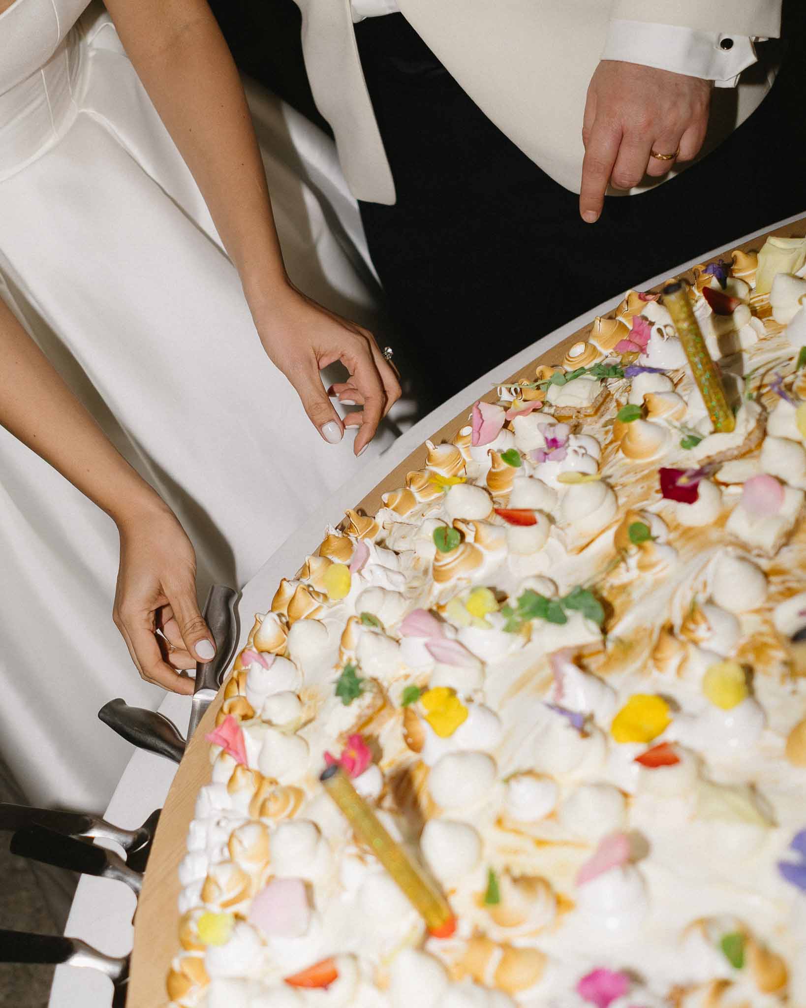 Overhead close-up of bride and groom cutting pavlova-style wedding cake topped with meringue rosettes and edible flowers