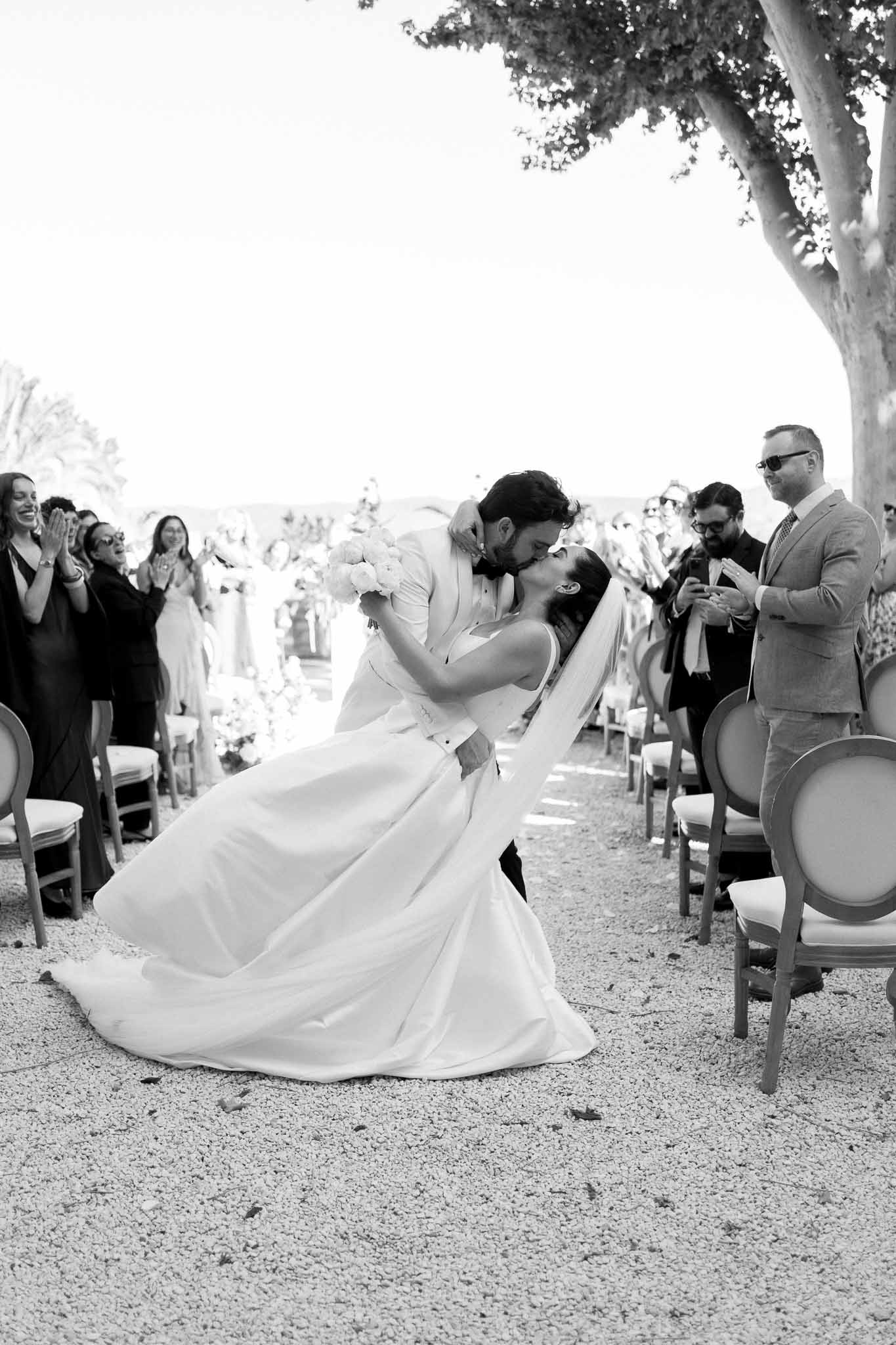 Black and white of groom dipping bride for kiss in ceremony aisle as guests clap on French Louis chairs