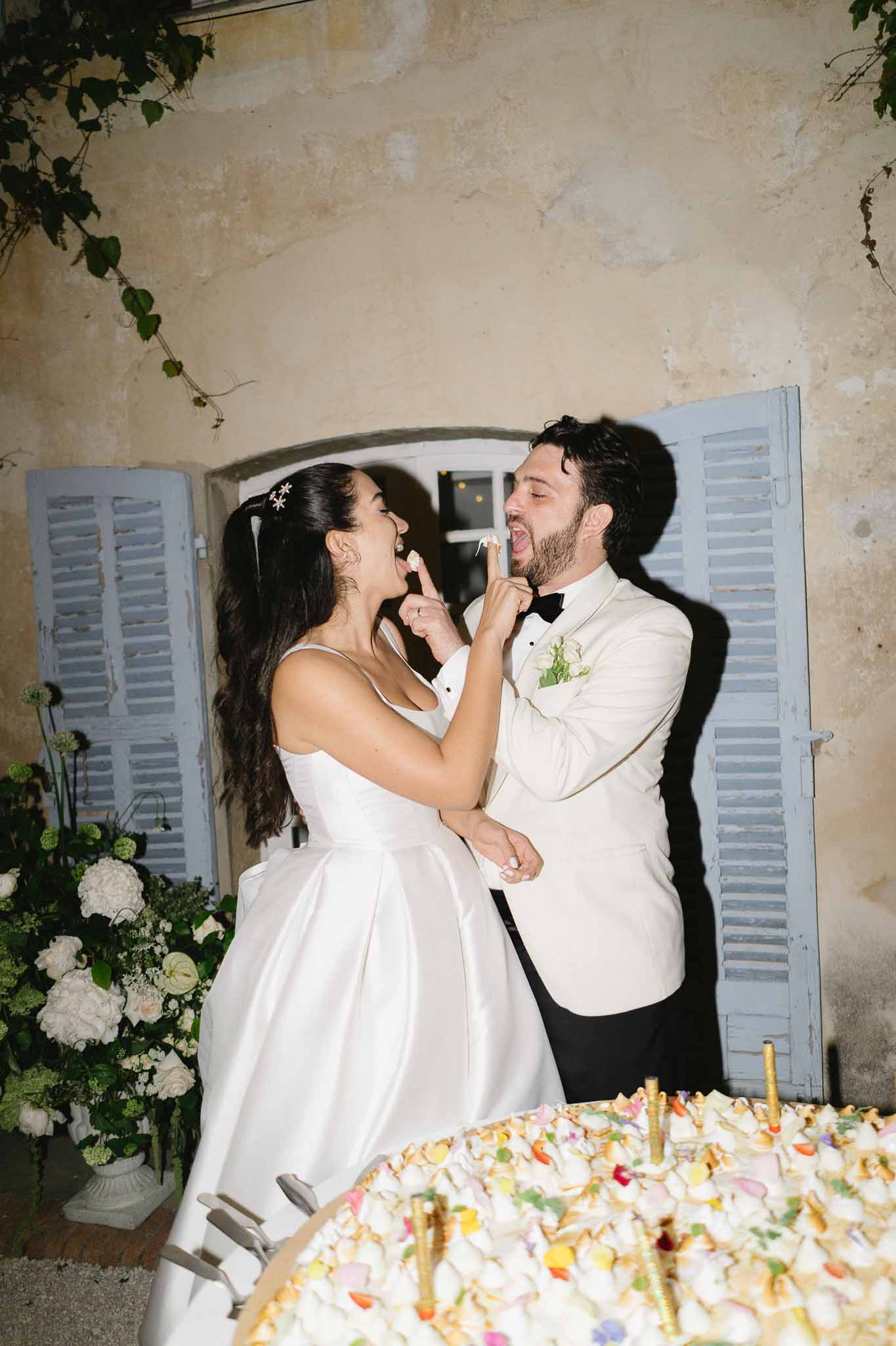 Bride and groom laughing while feeding each other cake at evening outdoor reception in front of chateau with blue shutters