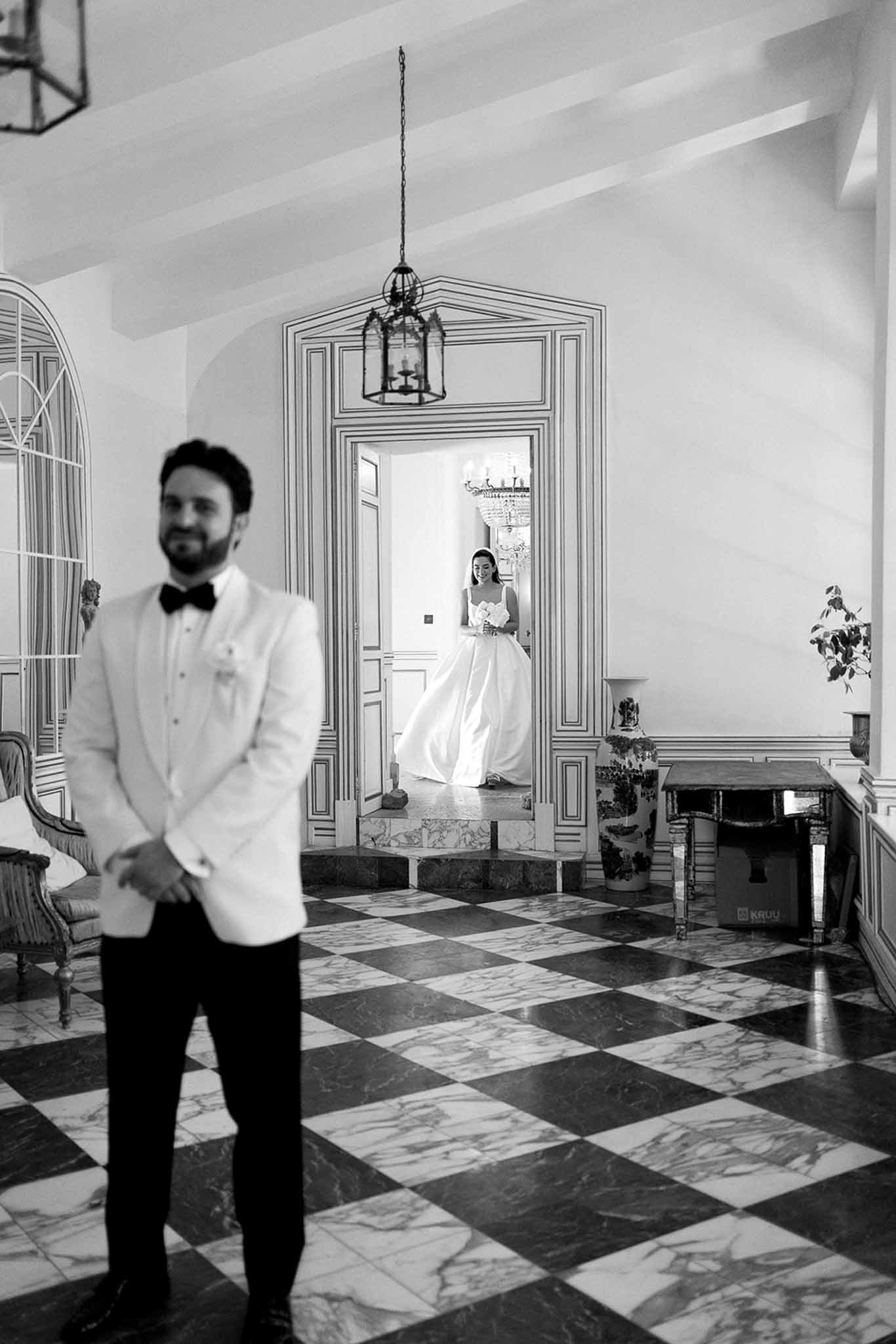 Black and white first look through ornate doorway on checkered marble floor with chandelier and armchairs