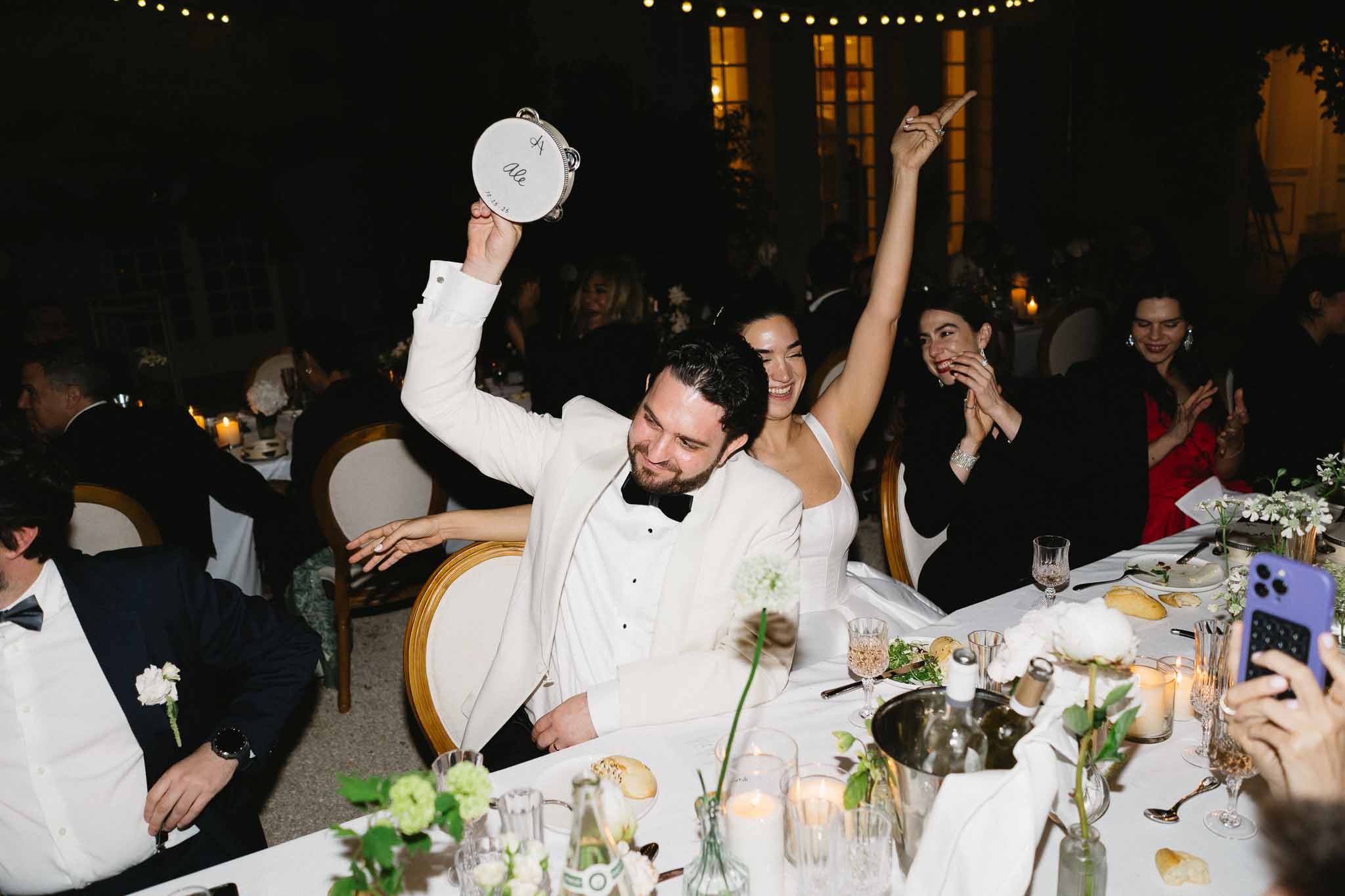 Bride and groom celebrating at outdoor reception head table with raised arms, guests laughing, string lights overhead