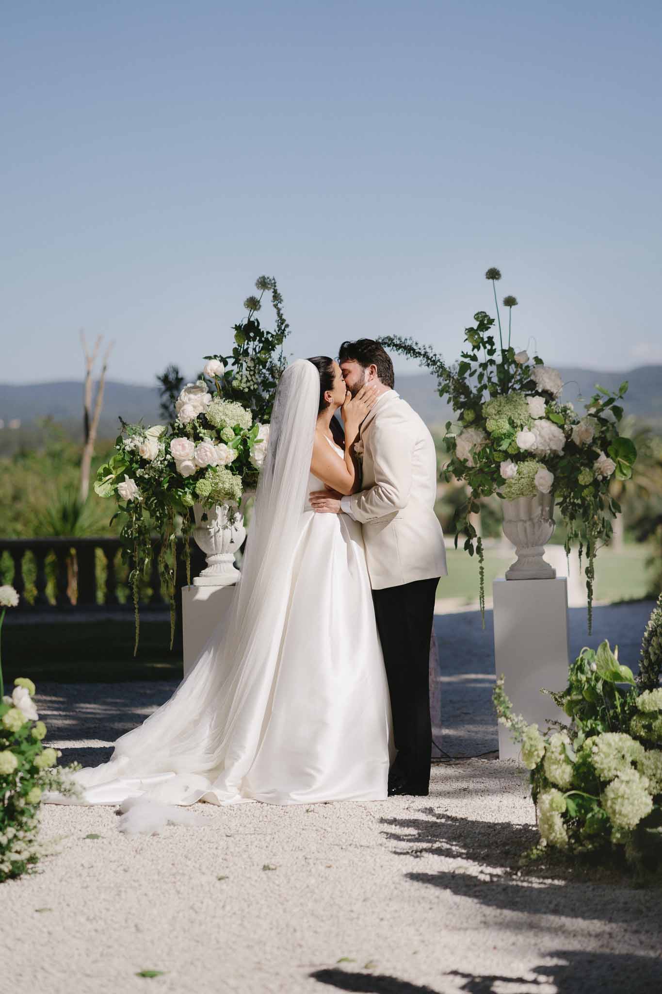 Bride and groom share first kiss before circular greenery arch flanked by white rose urns on plinths
