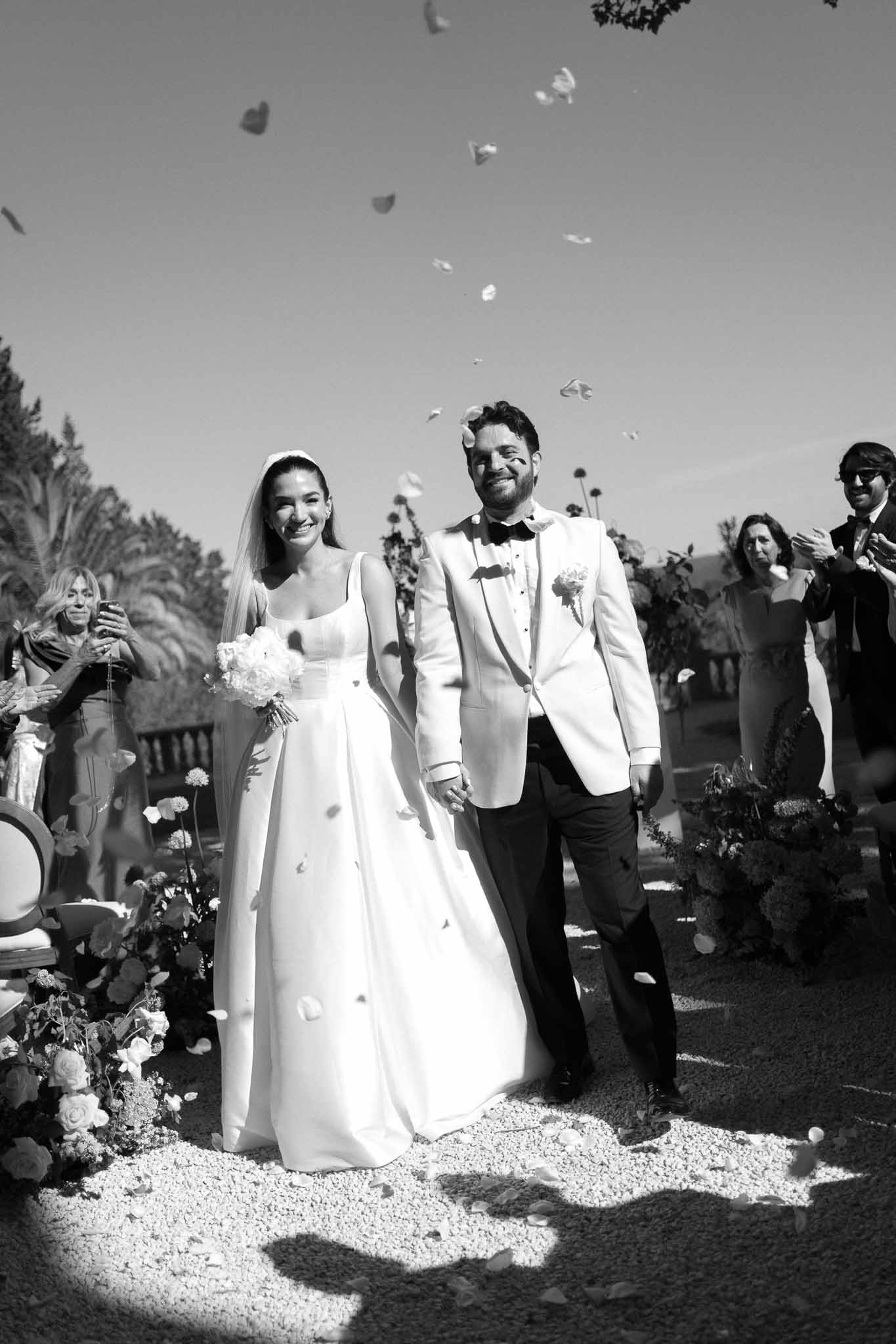Bride and groom walking down outdoor aisle with flower petals tossed by guests in black and white