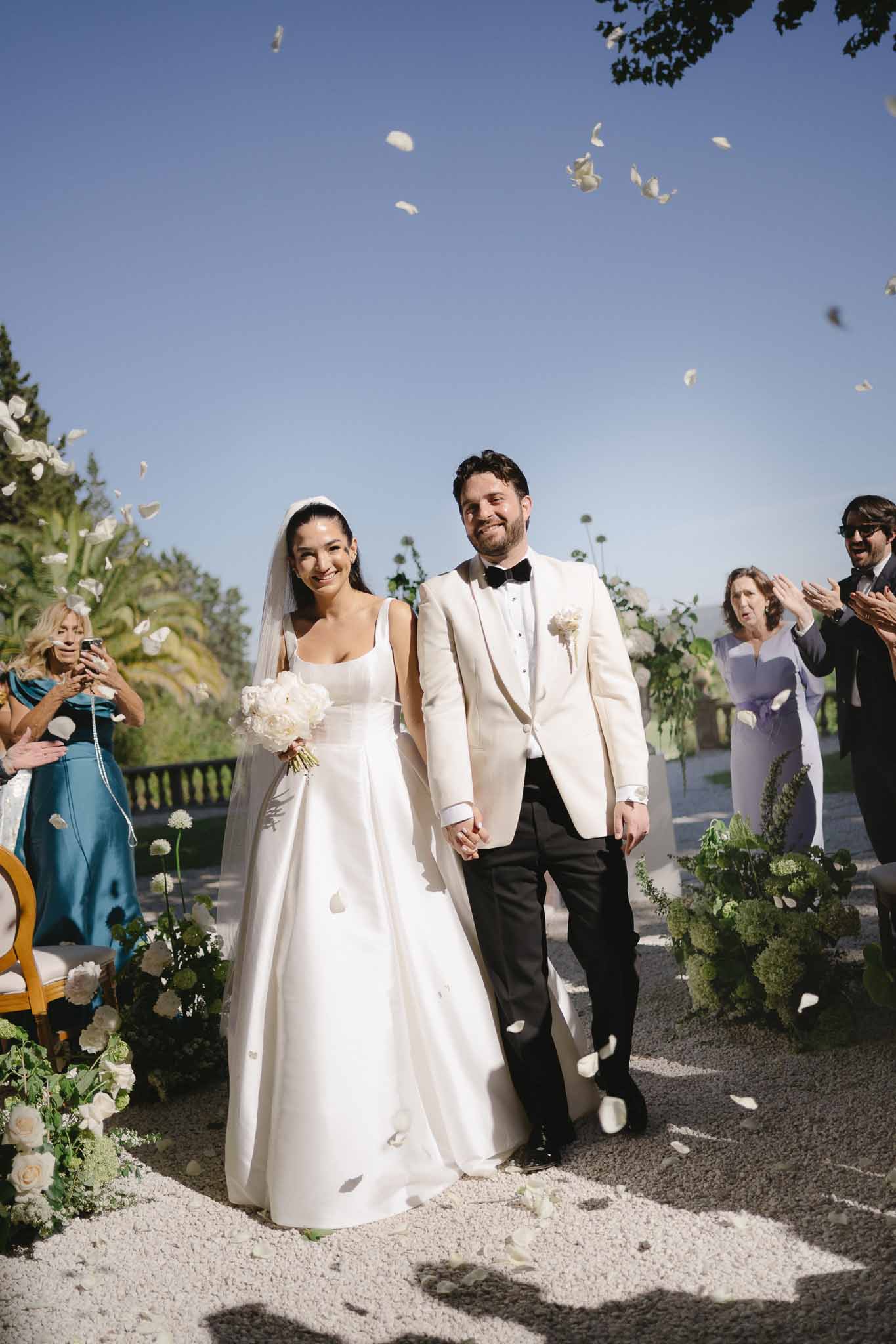 Bride and groom walk down aisle as guests toss white petals with white rose arrangements lining path
