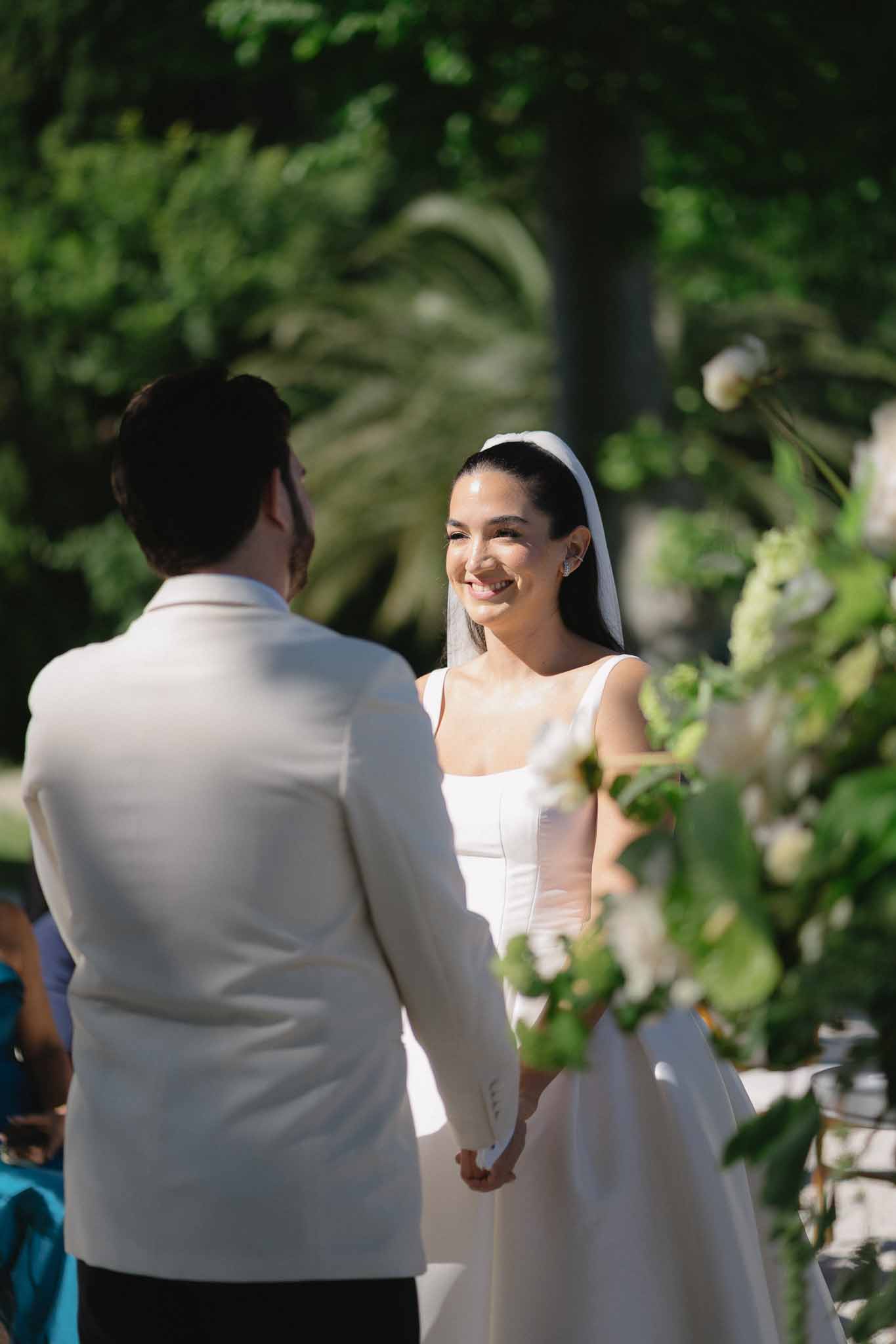 Bride and groom holding hands during outdoor garden ceremony with white floral arrangement and green foliage