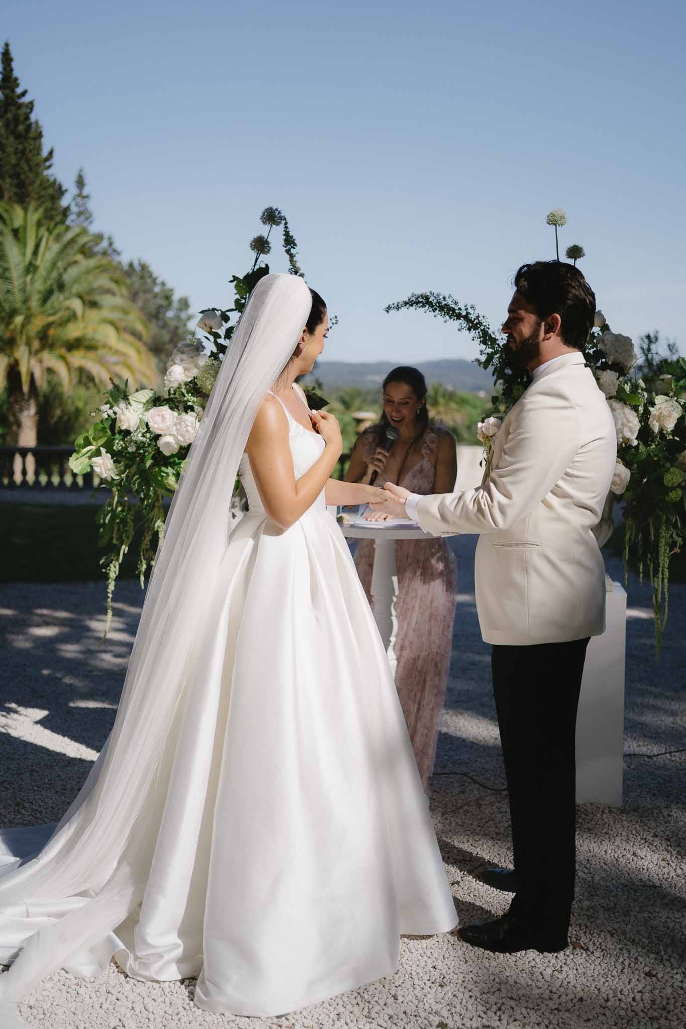 Couple exchanges vows under greenery hoop arch with hydrangeas as laughing officiant holds microphone