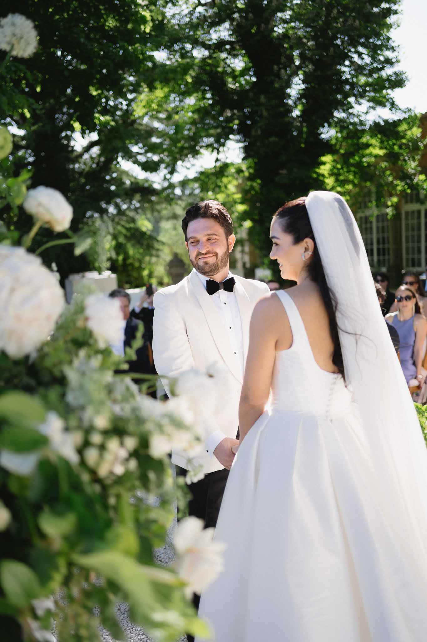 Couple faces each other at garden altar framed by tall white viburnum arrangements with veil trailing