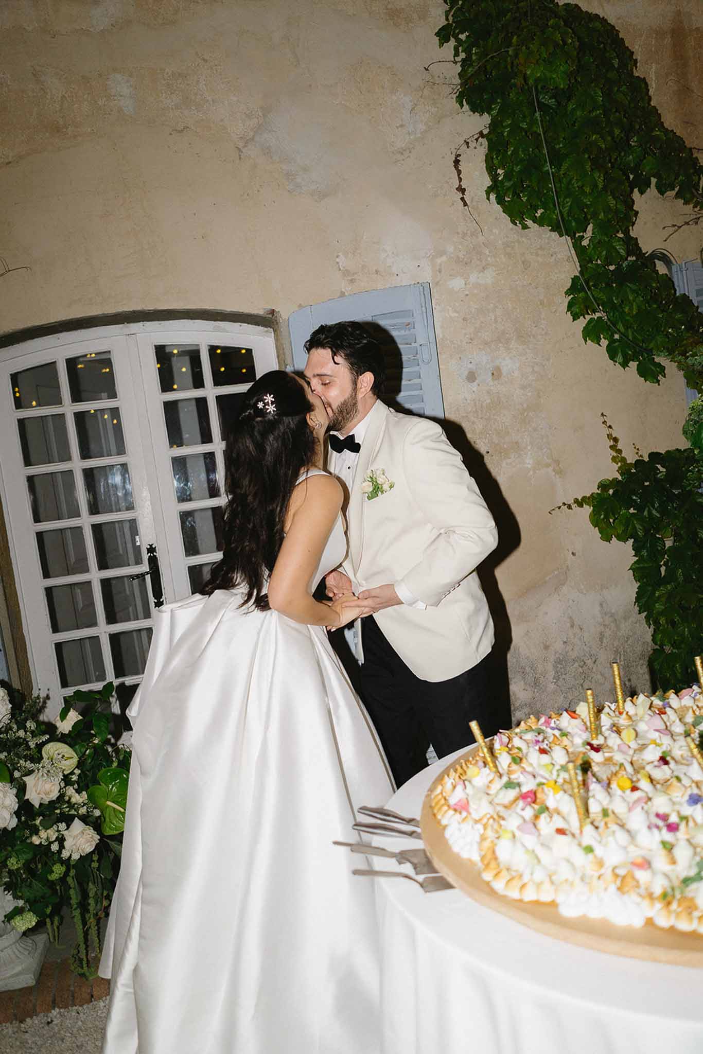 Bride and groom kissing during cake cutting at outdoor evening reception beside stone wall with ivy