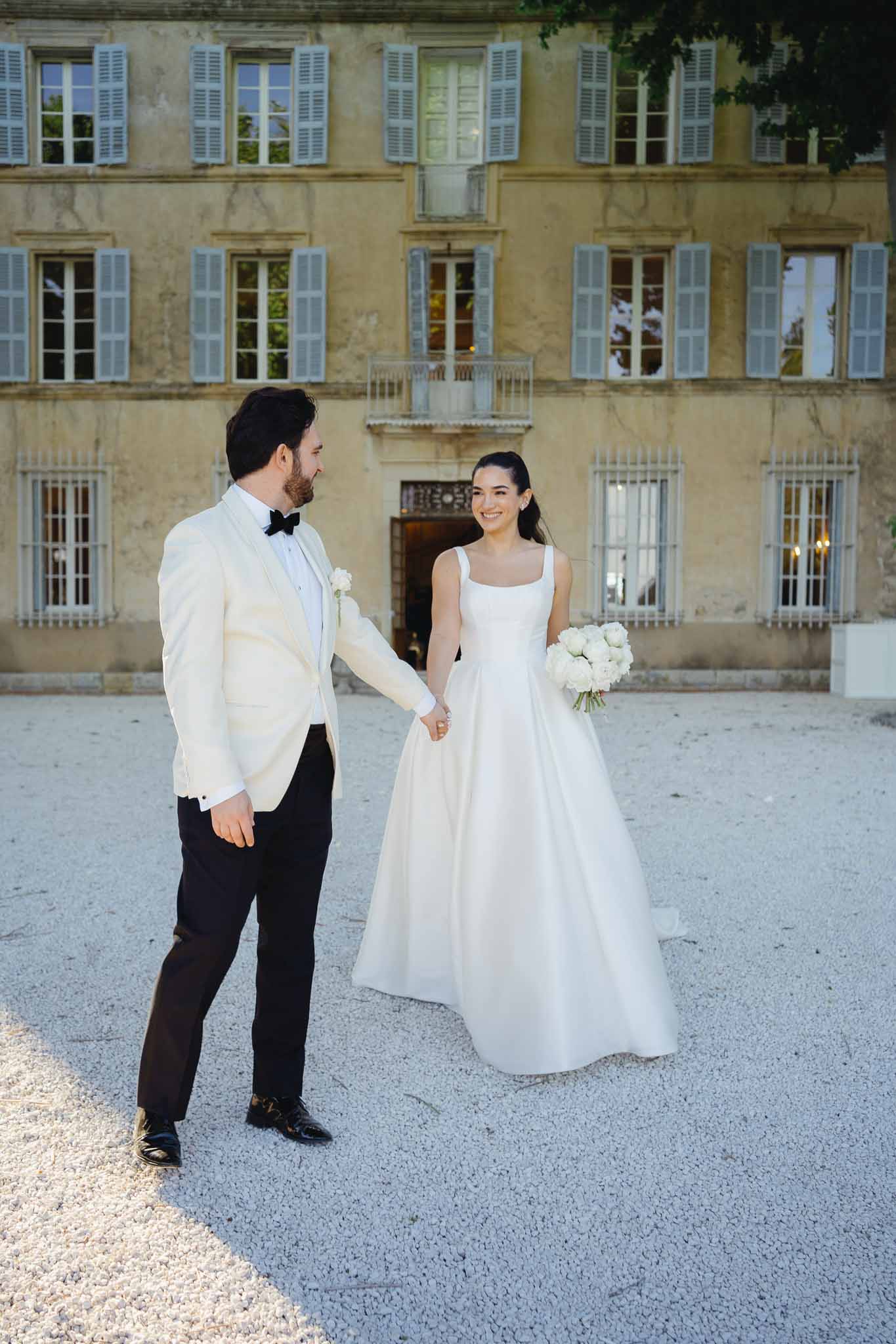 Bride in ivory ballgown and groom in ivory dinner jacket holding hands before ochre chateau facade