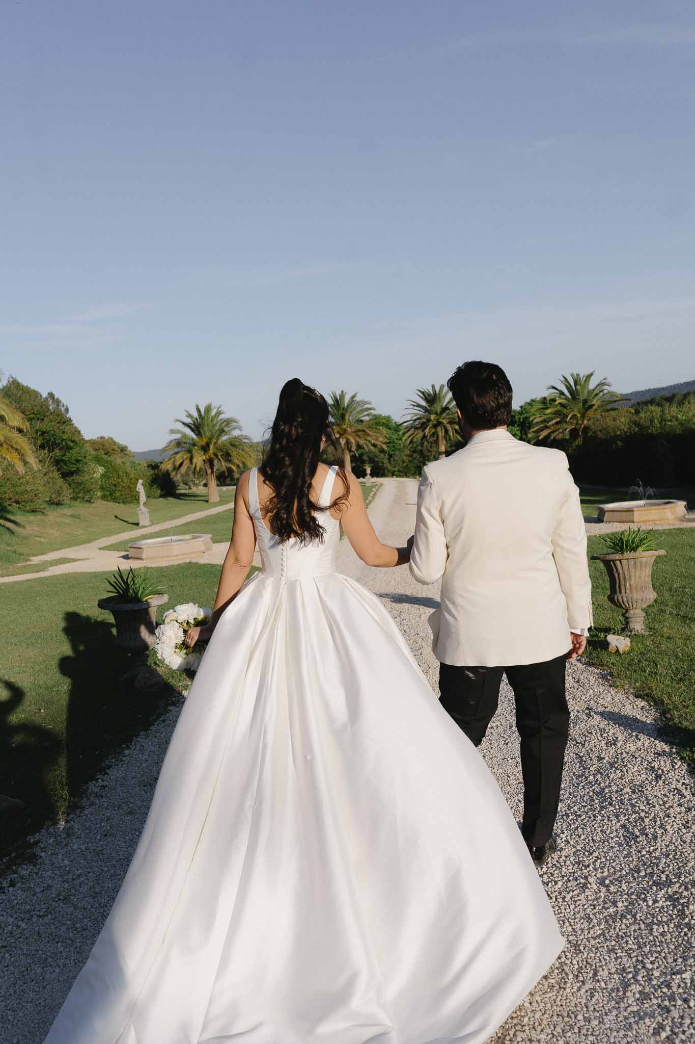 Couple from behind walking through formal garden with stone urns fountain basins and palm trees at Mediterranean estate
