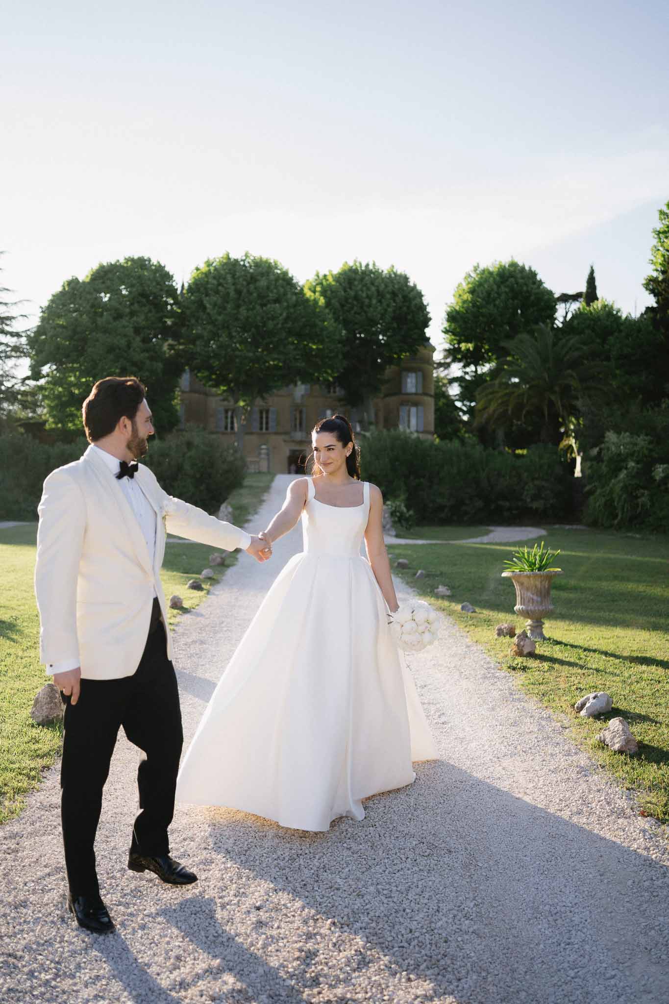 Bride in white ball gown and groom in white dinner jacket holding hands on chateau gravel driveway at sunset
