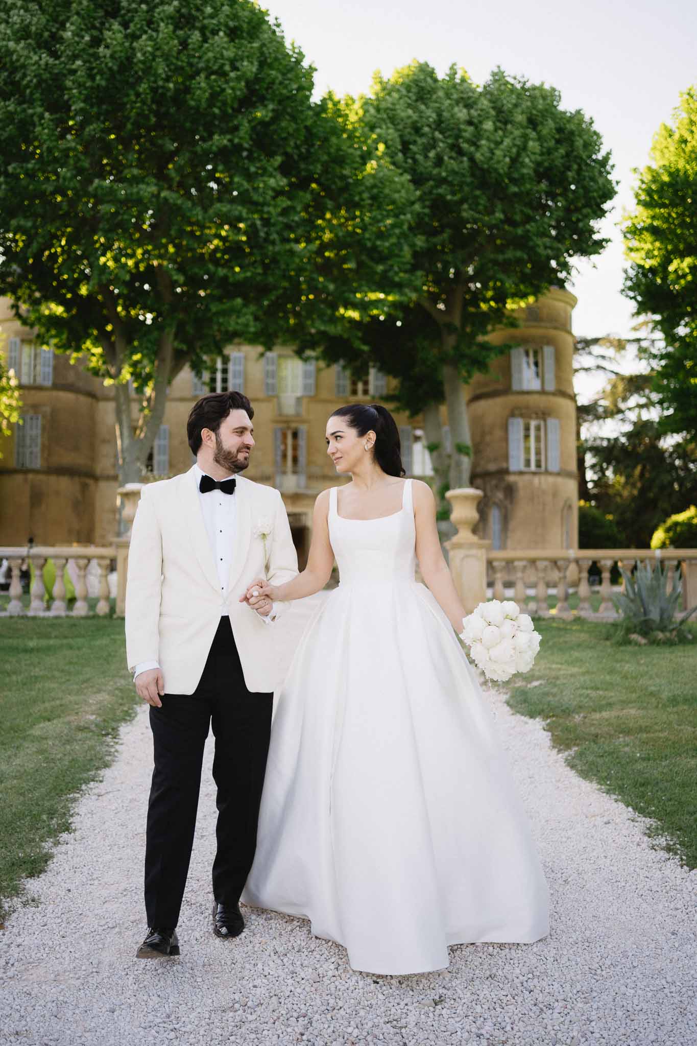Couple walking hand in hand before golden-stone chateau with tower, bride carrying white peony bouquet