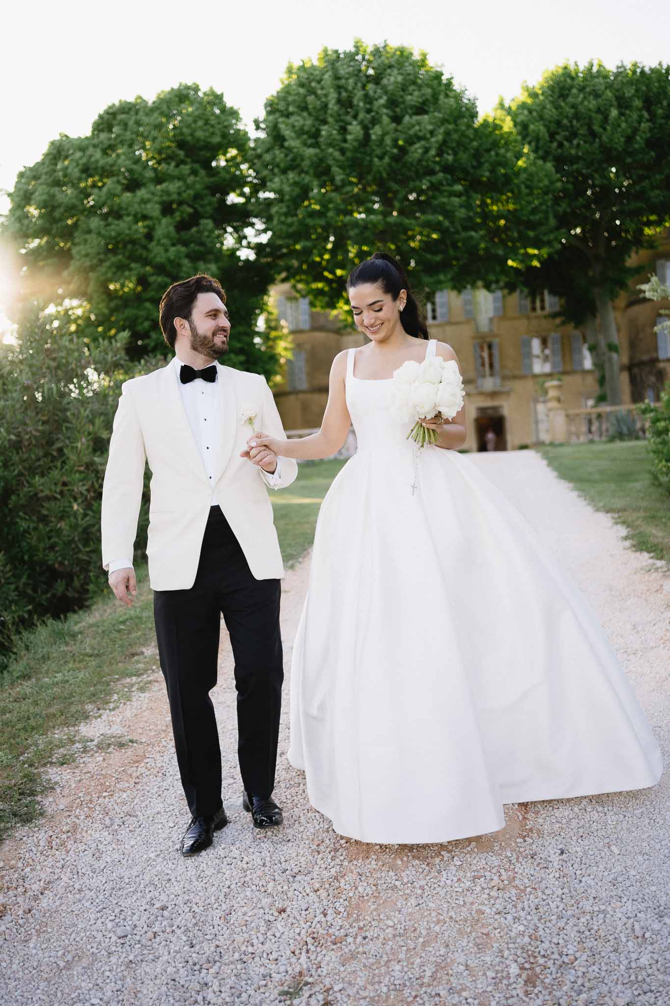 Bride in white ball gown and groom in white dinner jacket walking on gravel path in front of French chateau at golden hour