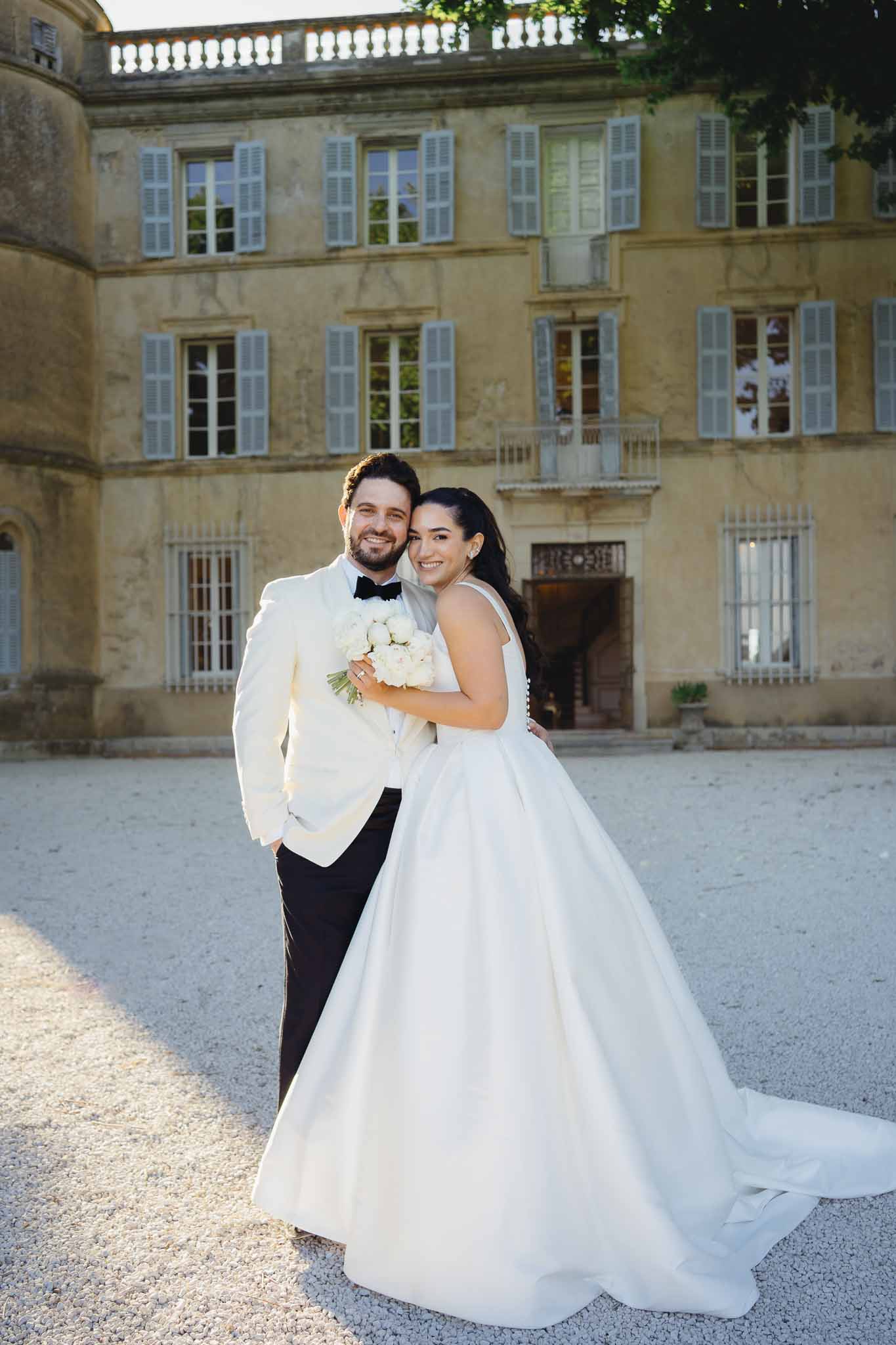 Bride in white ballgown with peony bouquet and groom in cream dinner jacket at chateau forecourt at golden hour
