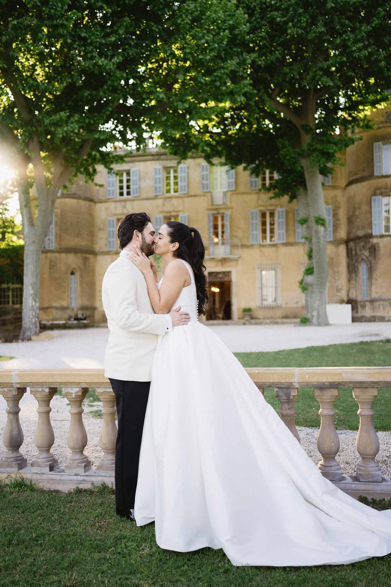 Bride and groom kissing at golden hour beside stone balustrade with French chateau in background
