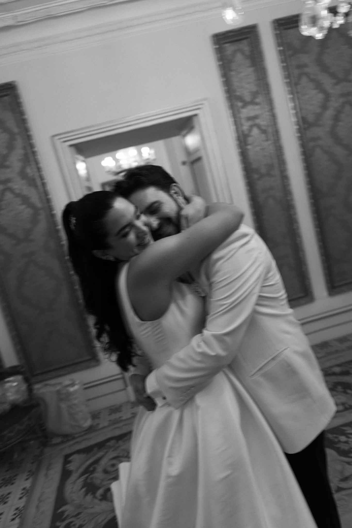 Bride and groom laughing together during first dance in an ornate ballroom with chandelier and tapestries