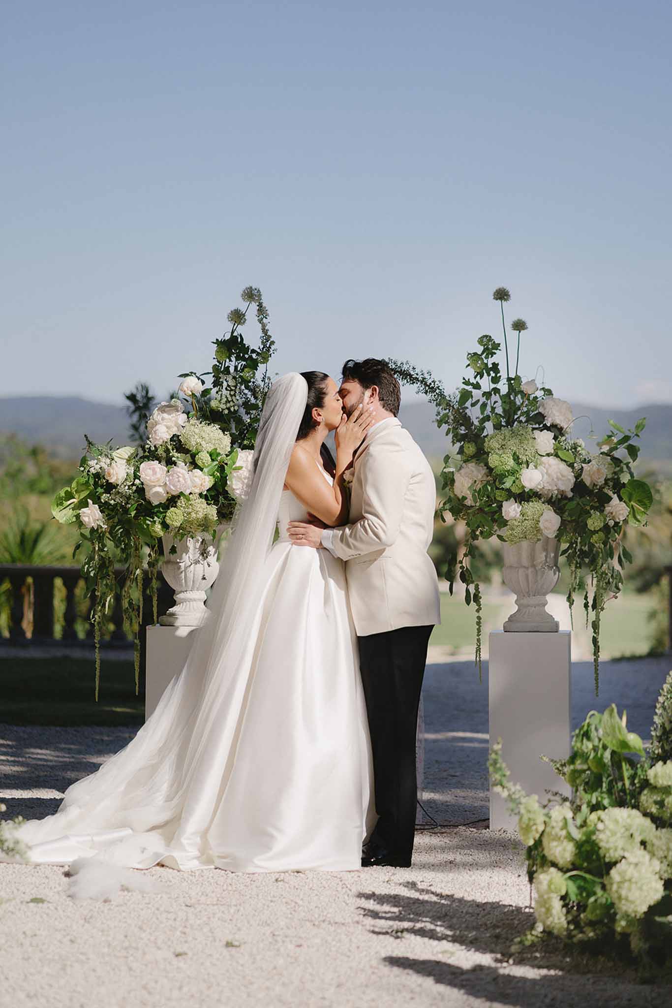 Couple shares first kiss between white rose urns on pedestals with green hydrangea on open-air terrace