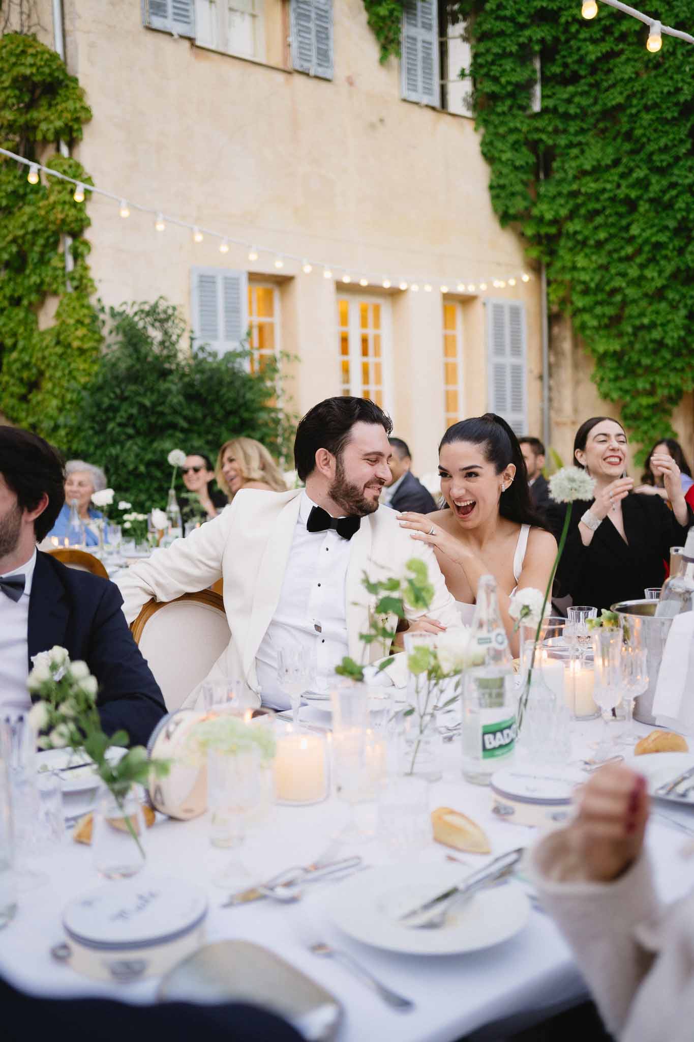 Bride and groom laughing at head table during outdoor evening reception in a chateau courtyard with string lights